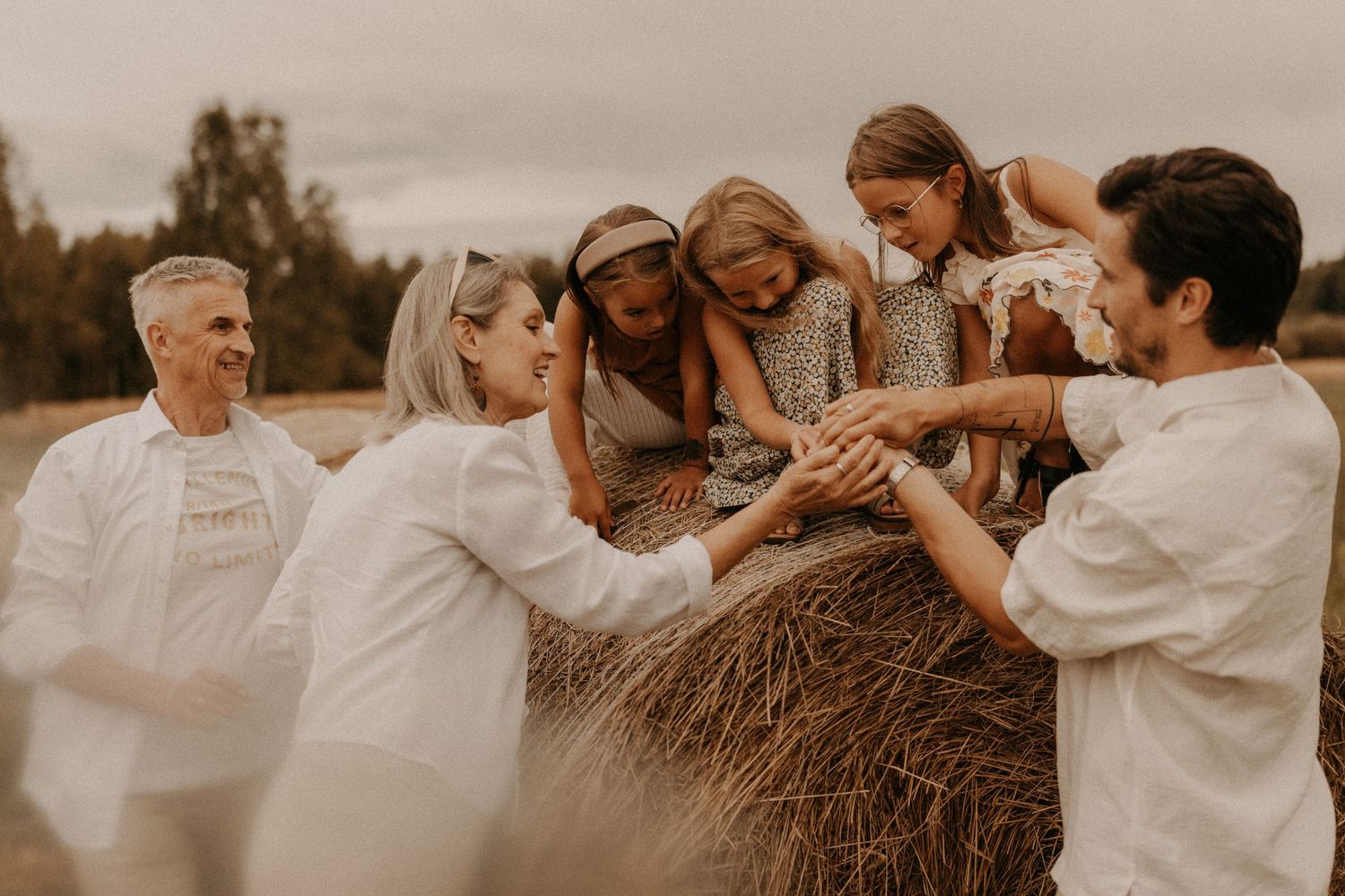 Family laughing together around a hay bale in a field, wearing white and neutral tones during an outdoor photo session.