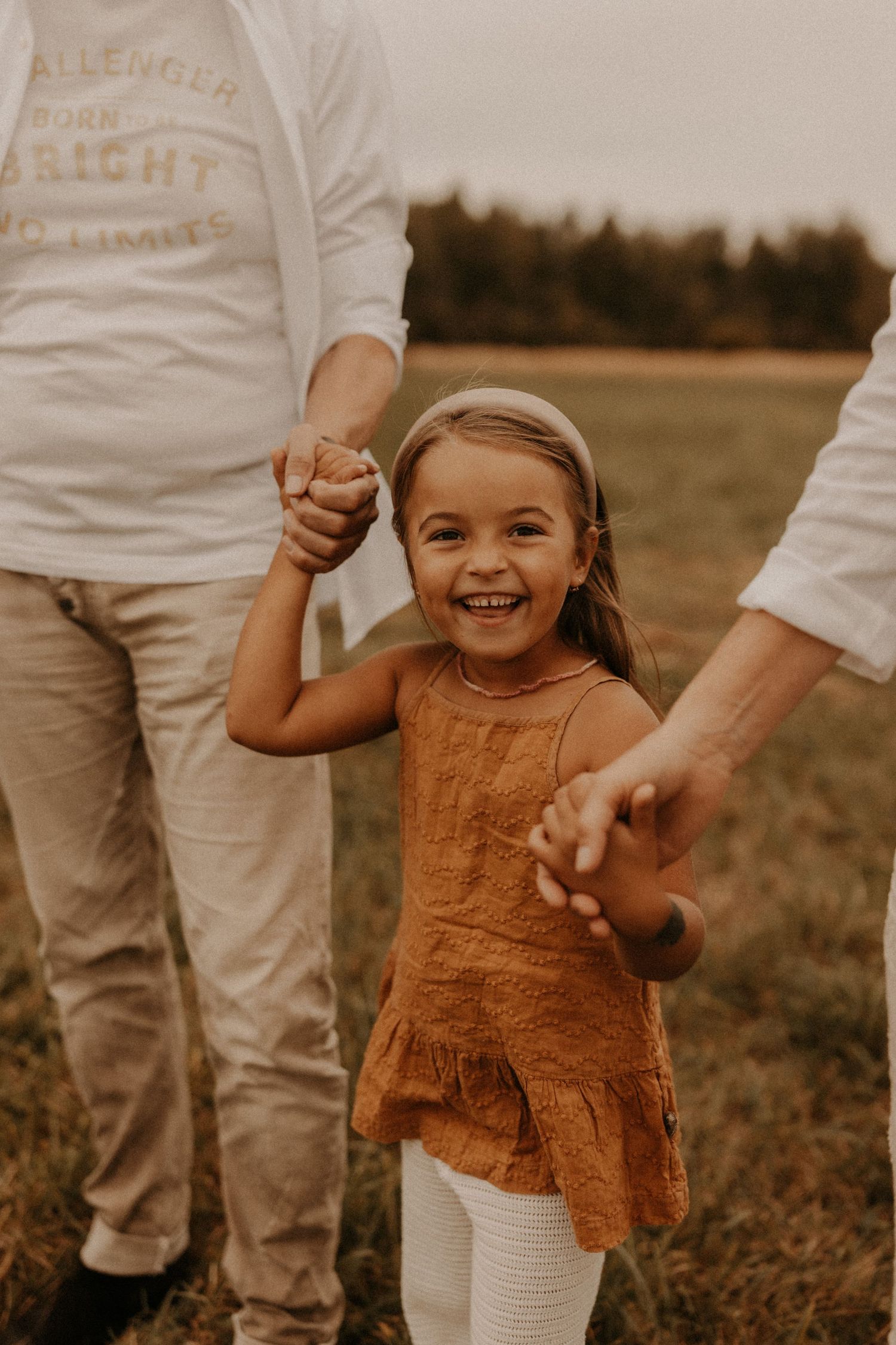 A joyful young girl in a rust dress holds hands with two adults in a green field, smiling brightly.