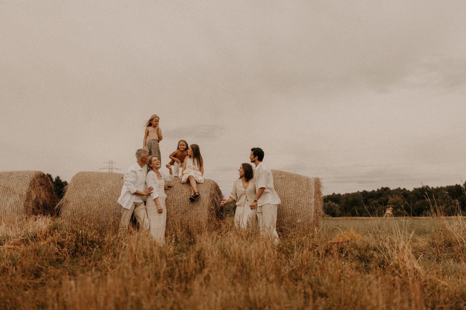 Family in white outfits posing on hay bales in a golden field under an overcast sky.