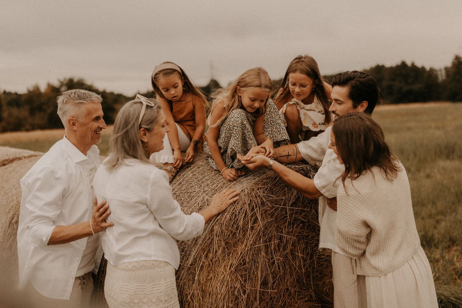 Family gathering around a hay bale in a field, dressed in white and neutral tones, sharing a joyful moment outdoors.