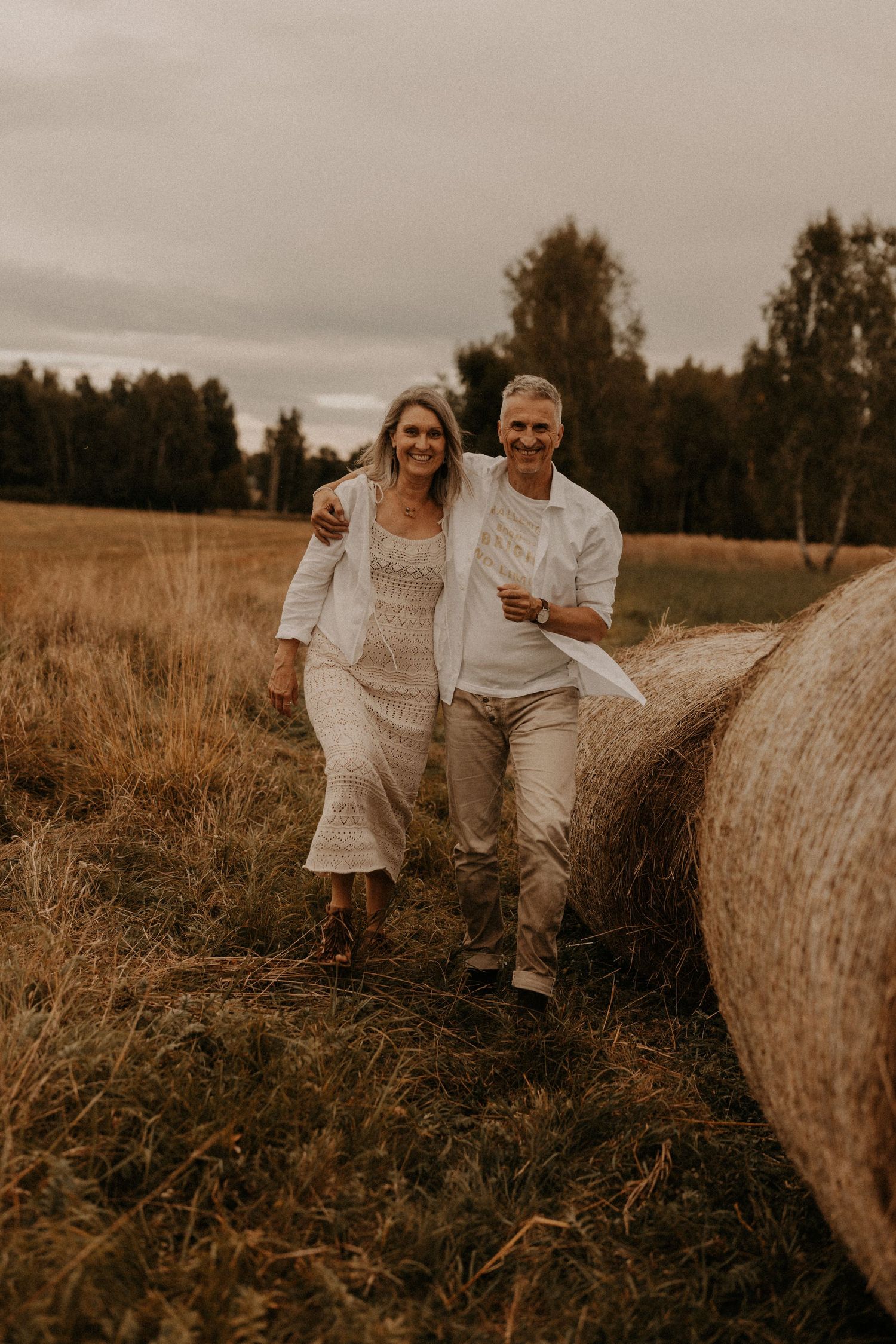 A smiling couple in white outfits walks through a golden field beside hay bales on a cloudy day.
