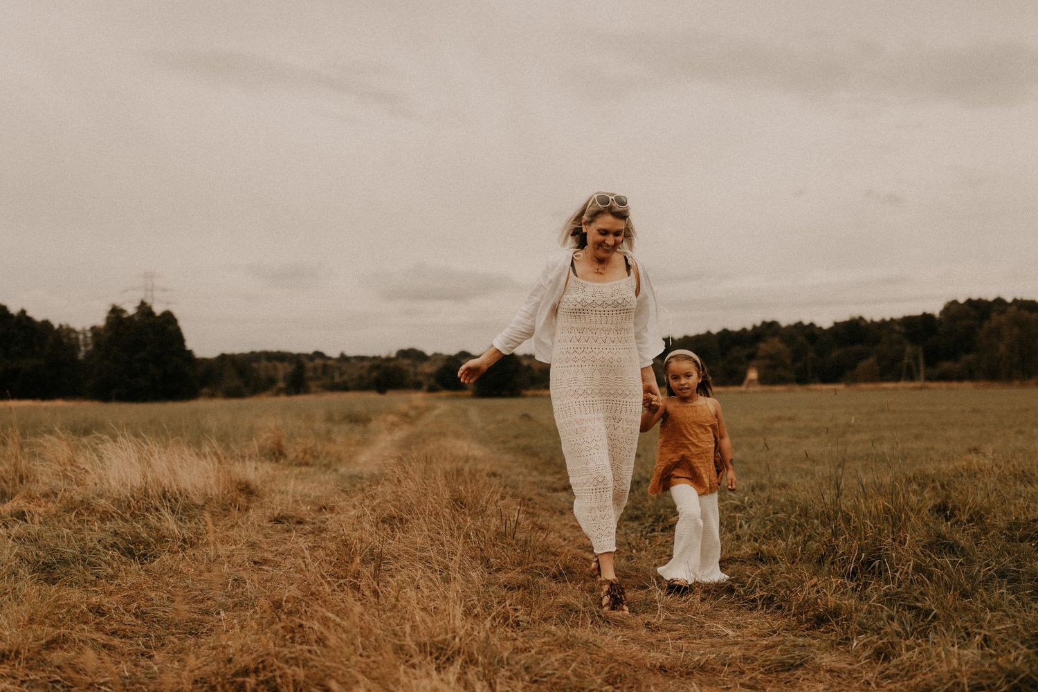 A mother in a white dress walks hand-in-hand with her toddler through an open autumn field.