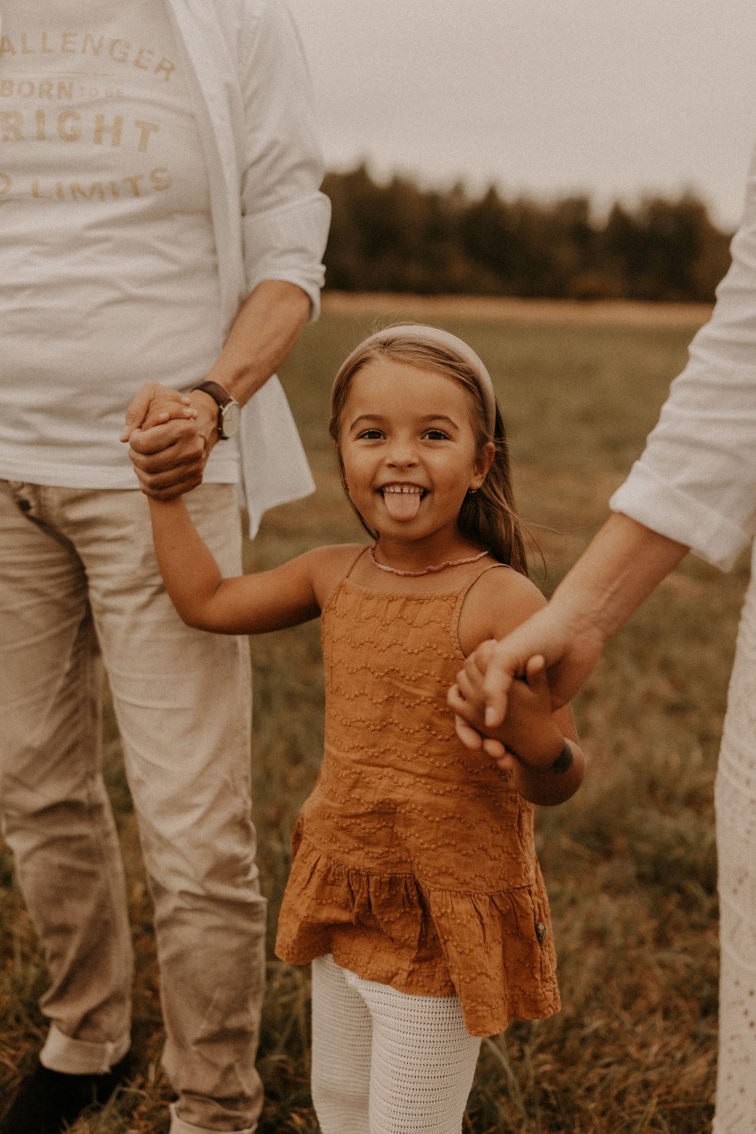 Young girl in orange dress laughs and sticks out tongue while holding hands with parents in a grassy field.
