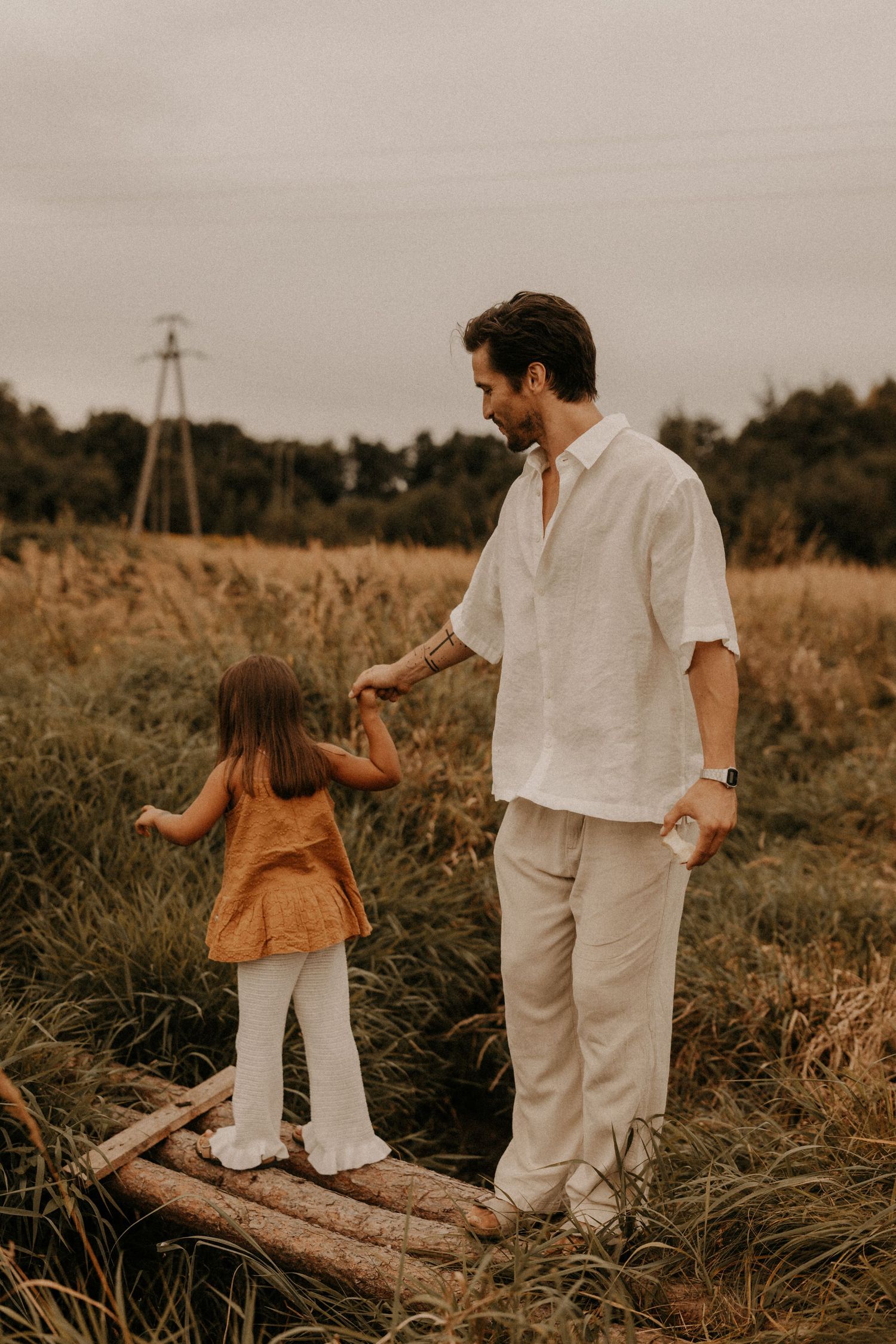 Father and daughter holding hands on a wooden log bridge in a golden field, wearing linen outfits.
