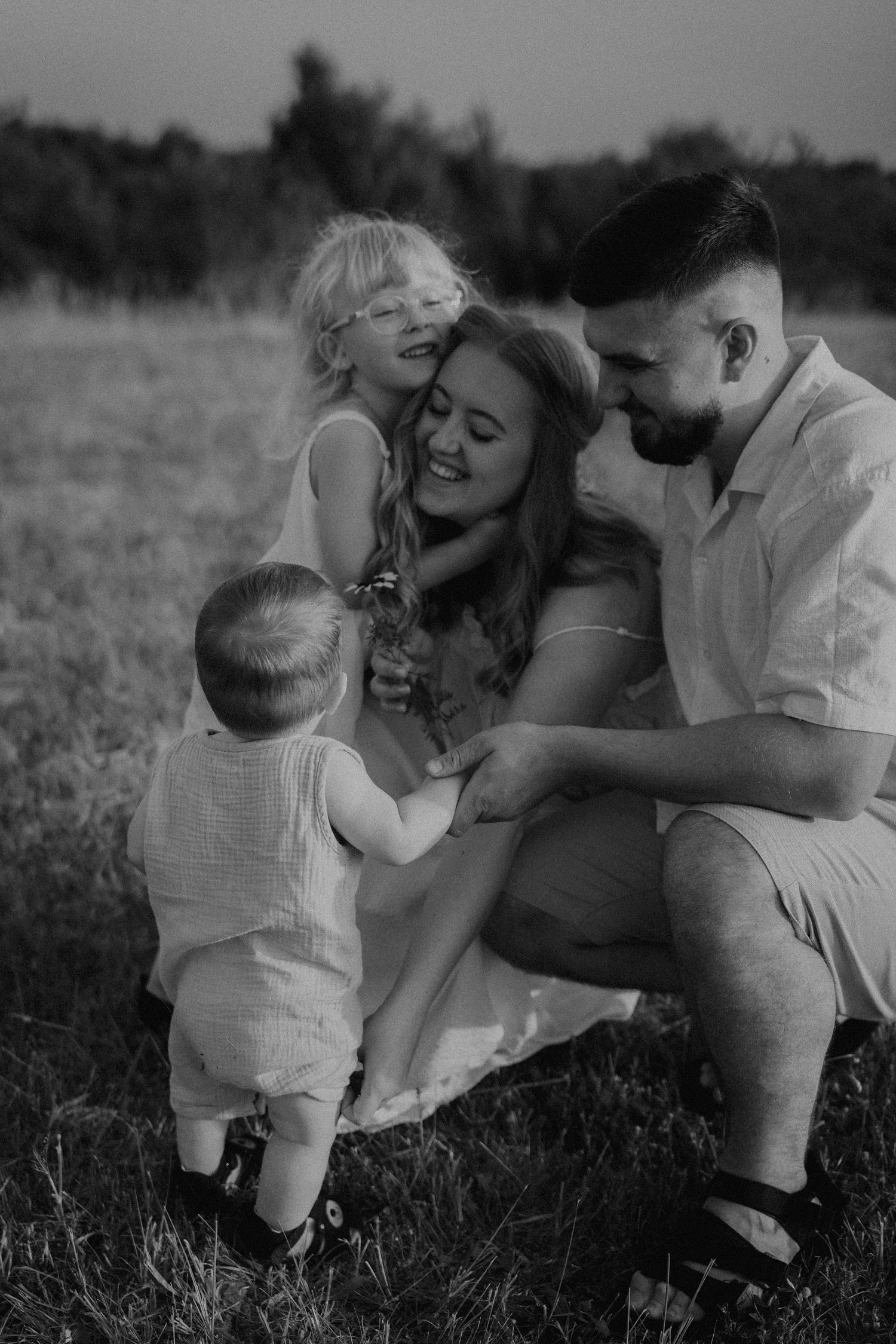 Black and white family photo of parents with two young children embracing in an outdoor field at dusk. sesje zdjęciowe Rybnik, Wodzisław Ślą