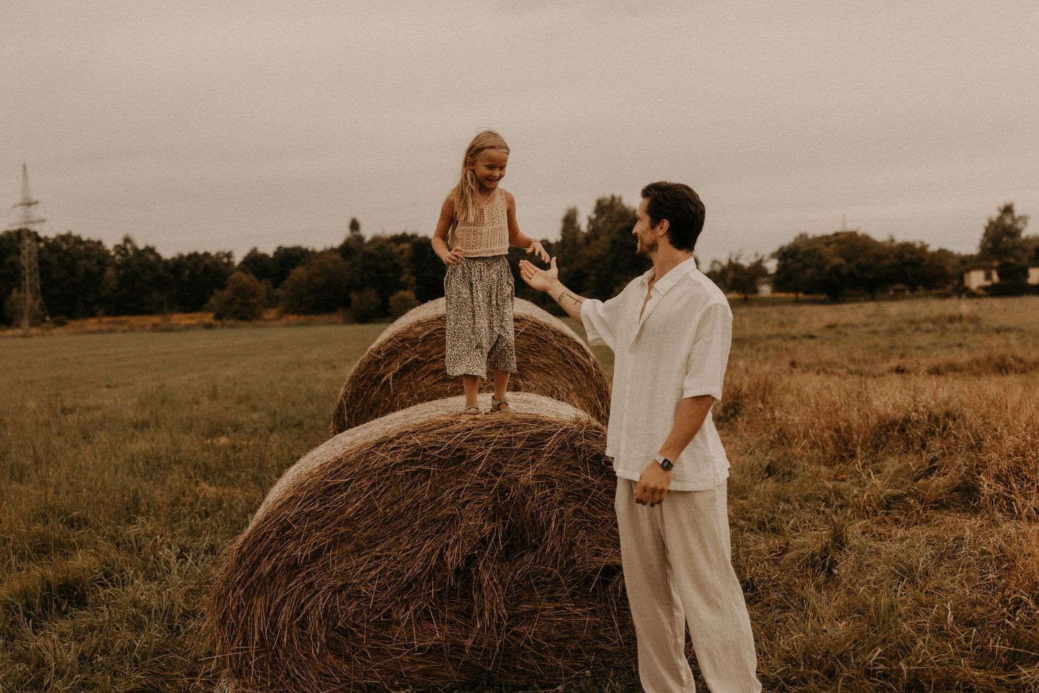A man helps a woman standing on a hay bale in a golden field, sharing a romantic countryside moment.