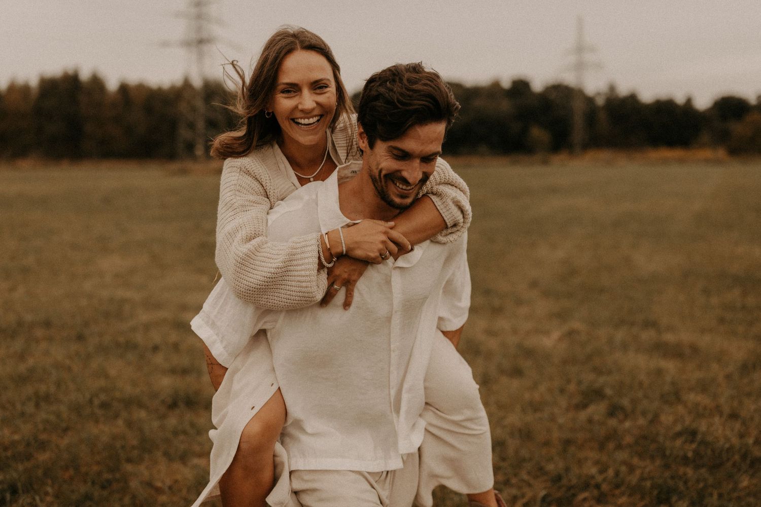 A joyful couple laughs together in a green field, the man giving the woman a piggyback ride in matching white outfits.