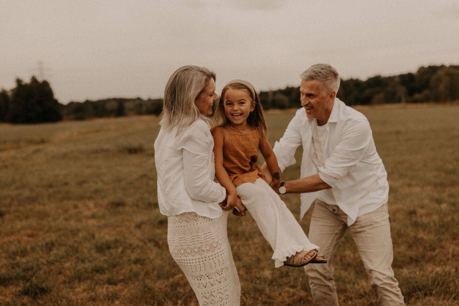A happy family with two adults and a child playing in a green field, dressed in white and brown tones.