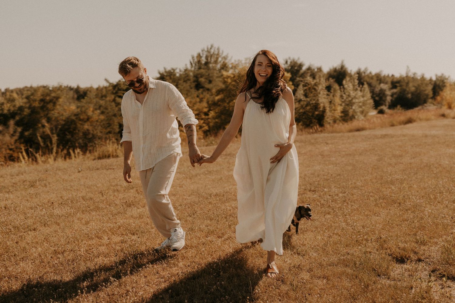 A pregnant woman in a white dress holds hands with a man walking through a sunny golden meadow. sesje zdjęciowe Rybnik, Wodzisław Śląski
