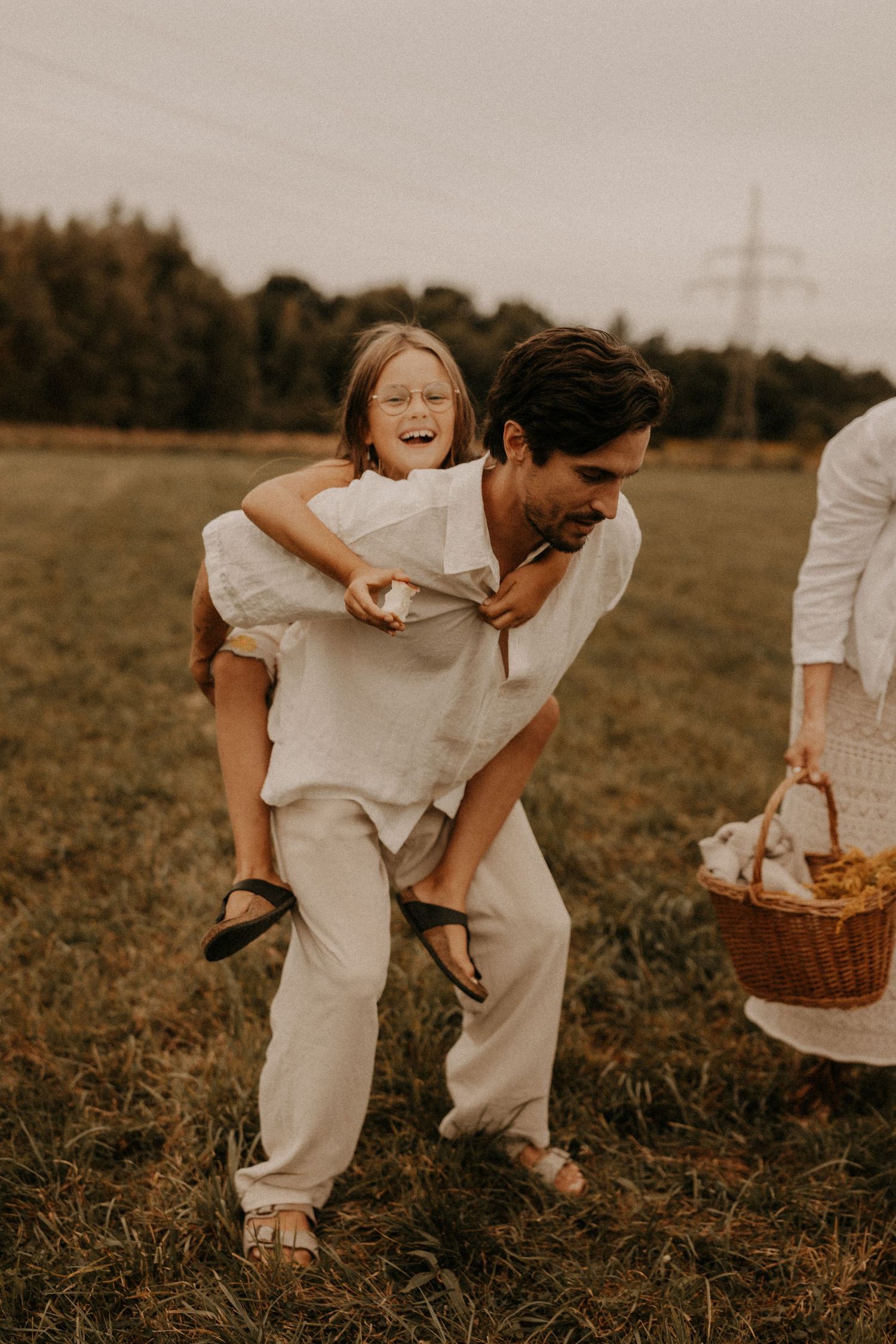 A man gives a laughing child a piggyback ride in a grassy field, while another person carries a wicker basket nearby.