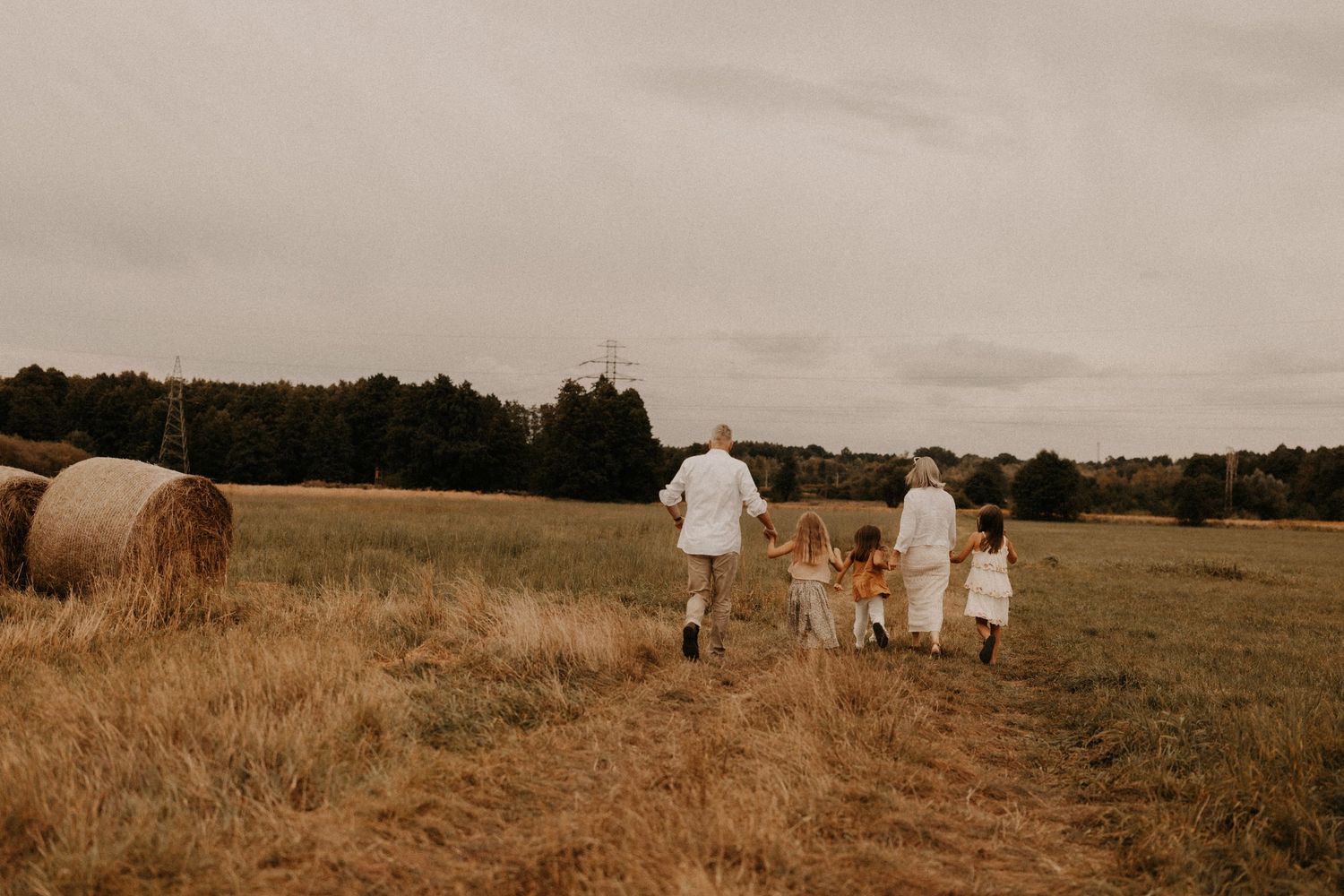 Family walking through golden hay field with round bale, trees in background on overcast day.