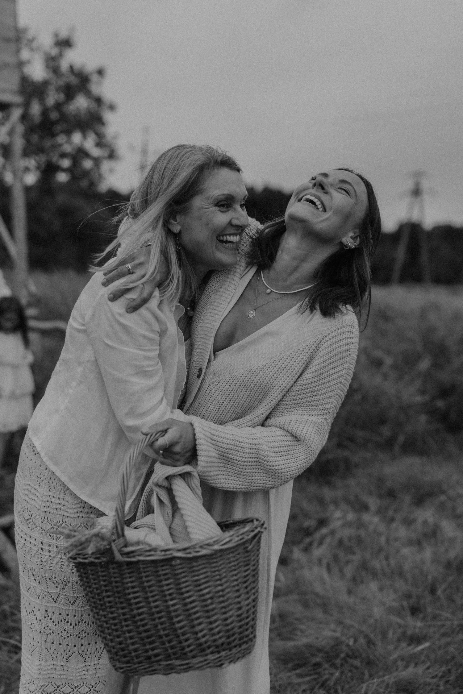 Two women laughing and embracing outdoors, holding a wicker basket in a black and white photo.
