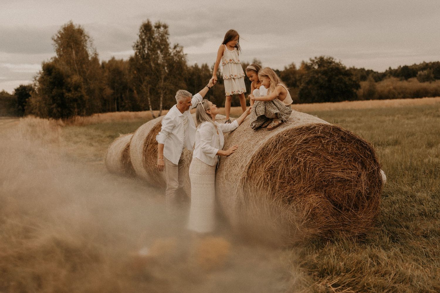 Family posing on hay bales in a golden field, children climbing while parents assist, rustic outdoor autumn setting.