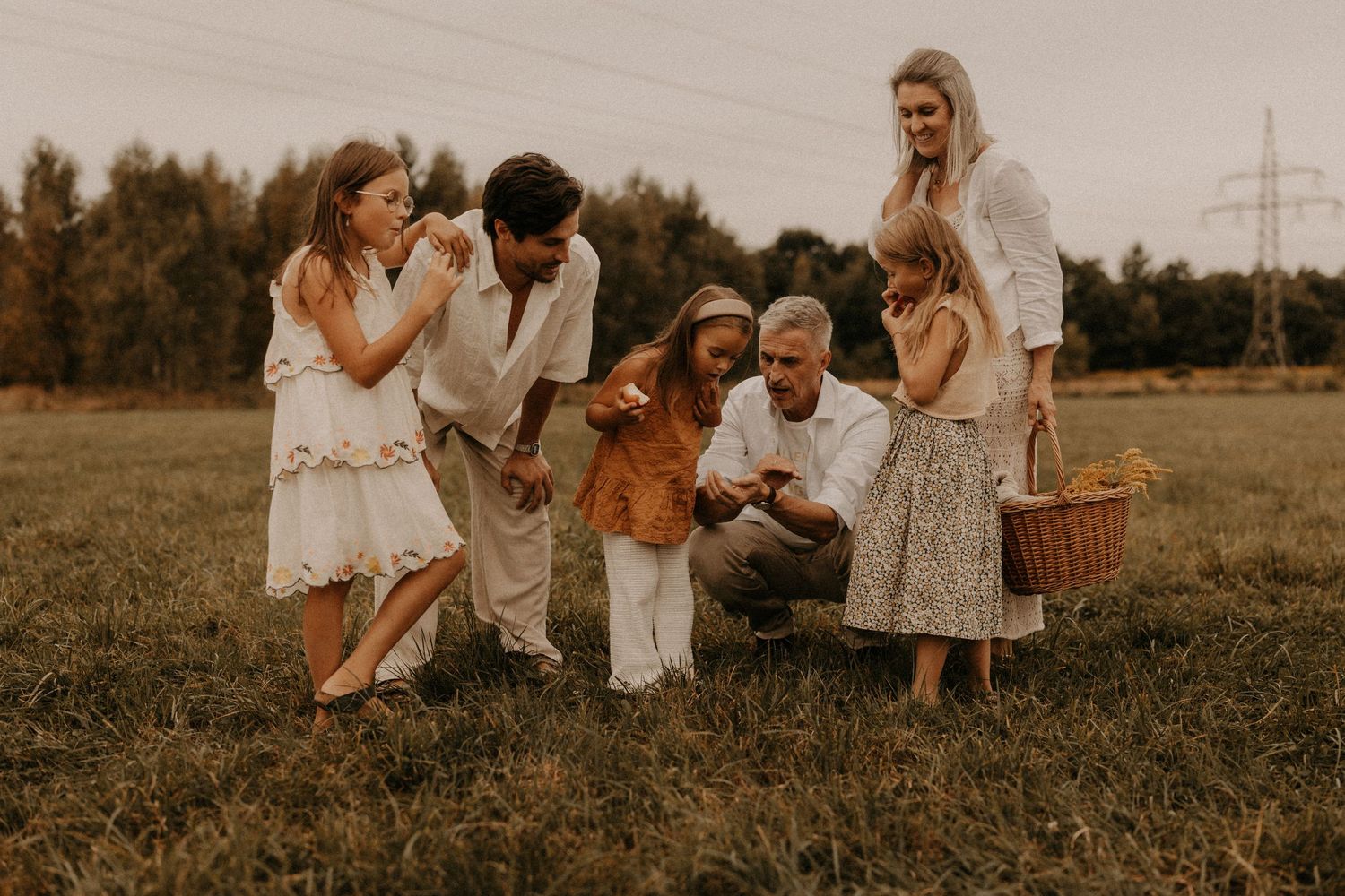 A warm family gathering in a field, with parents and children in earthy tones examining something together near a wicker basket.