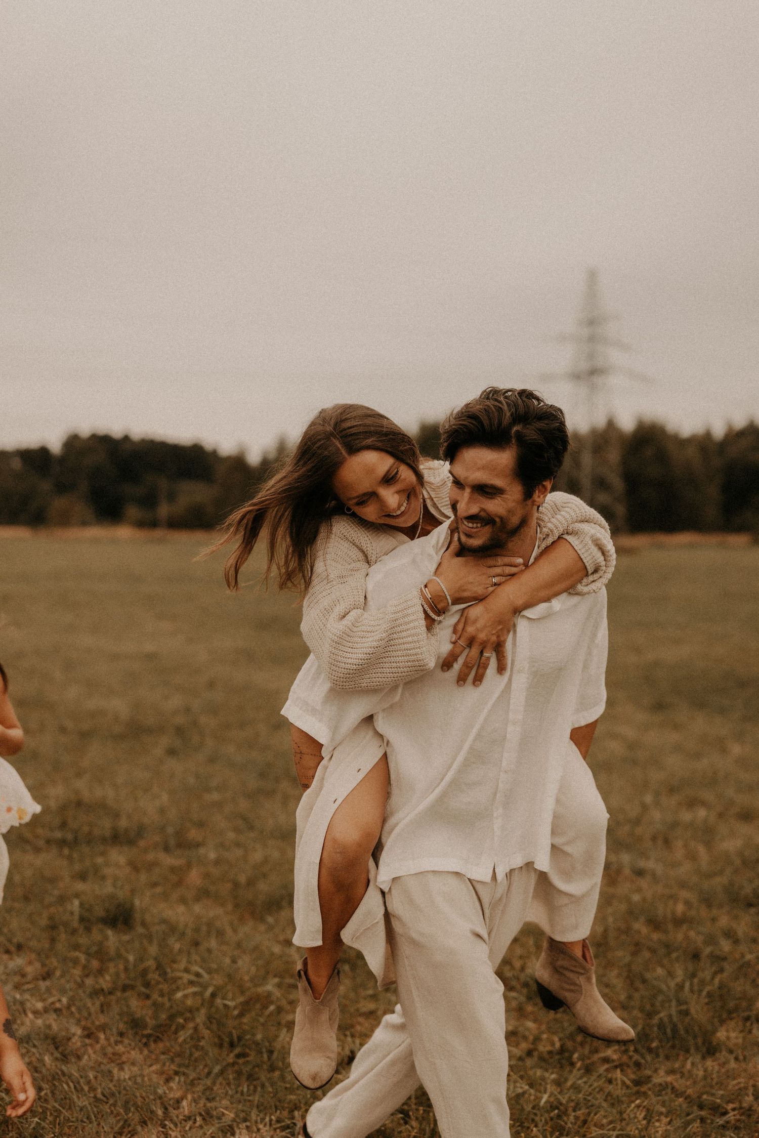 A joyful couple in white outfits share a piggyback ride in a green field, laughing together on a cloudy day.