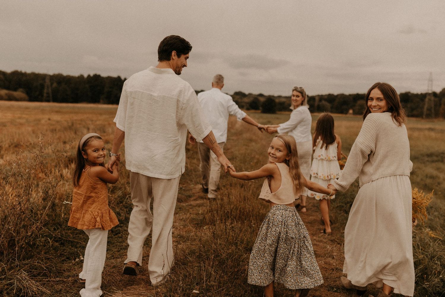 Happy family with children dancing in a circle on a field path, dressed in white and earth tones, outdoor portrait.