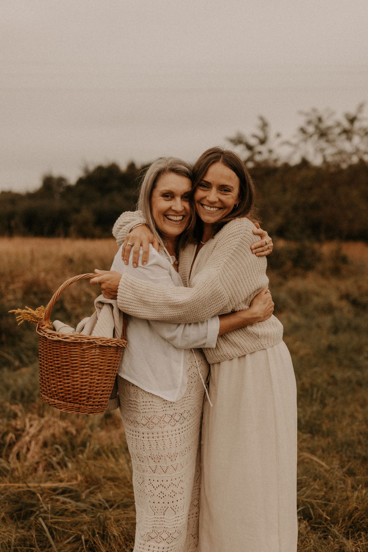 Two smiling women hugging in a field, one holding a wicker basket, wearing cozy neutral outfits on an overcast day.