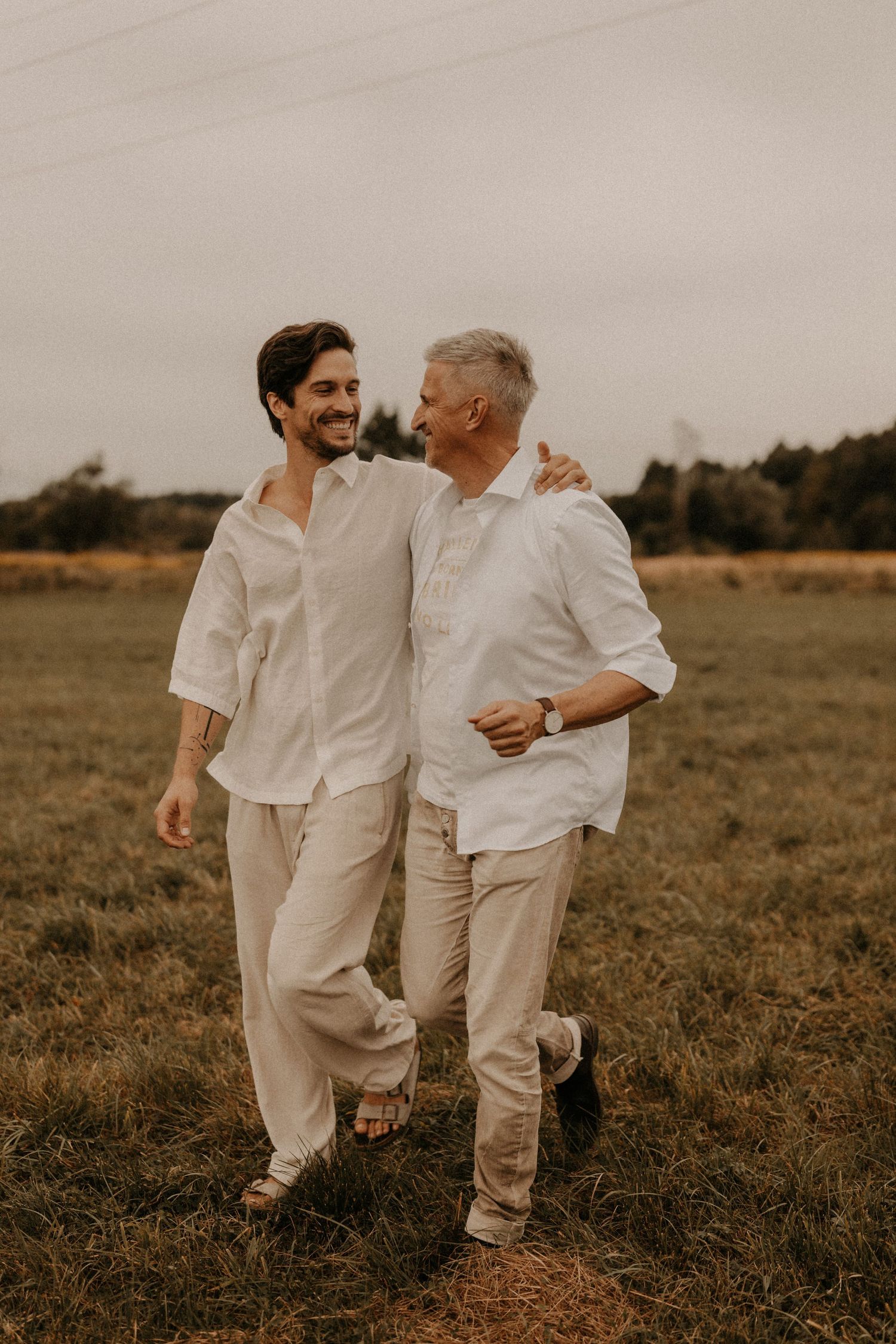 Two men in white linen shirts walk arm in arm through a field, smiling warmly at each other.