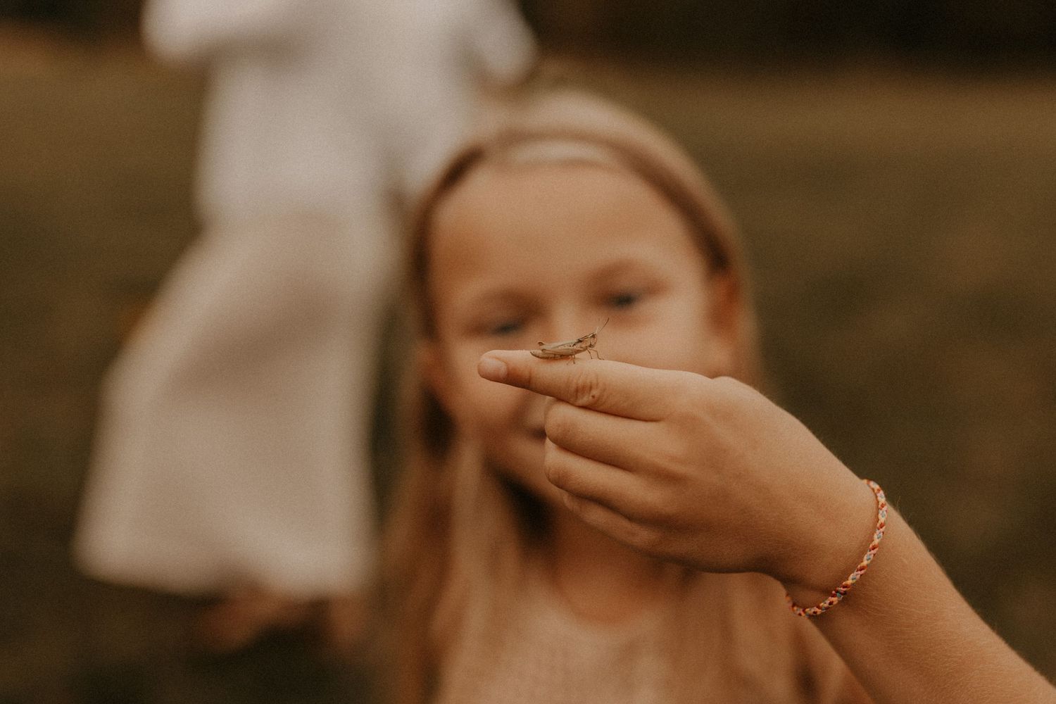 Young blonde girl covering her mouth with a hand wearing a gold bracelet outdoors.