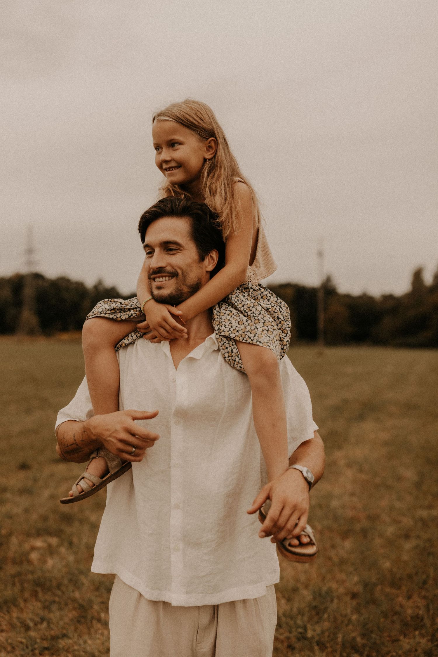 A smiling man carries a happy young blonde girl on his shoulders in a green field on a cloudy day.