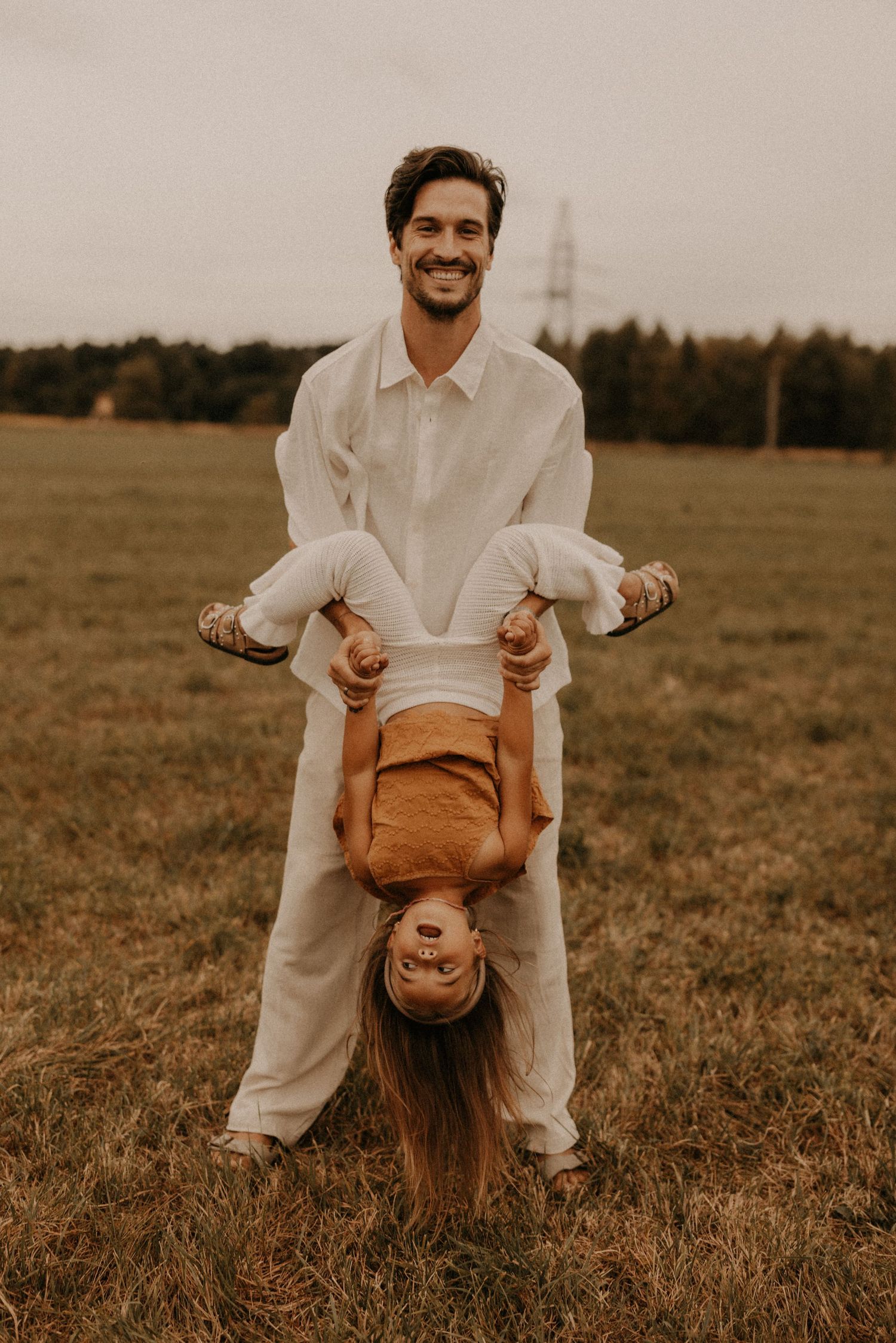 Smiling man in white outfit playfully holding a child upside down in a green field with trees in the background.