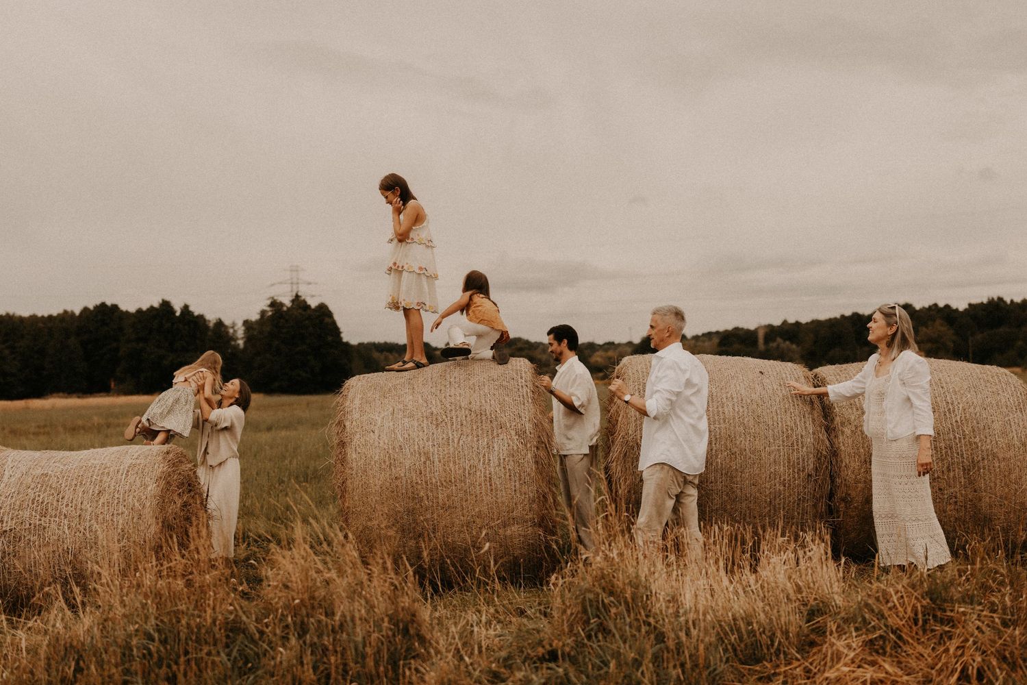 Family posing on large hay bales in a golden field at dusk, wearing neutral tones in a rustic outdoor setting.
