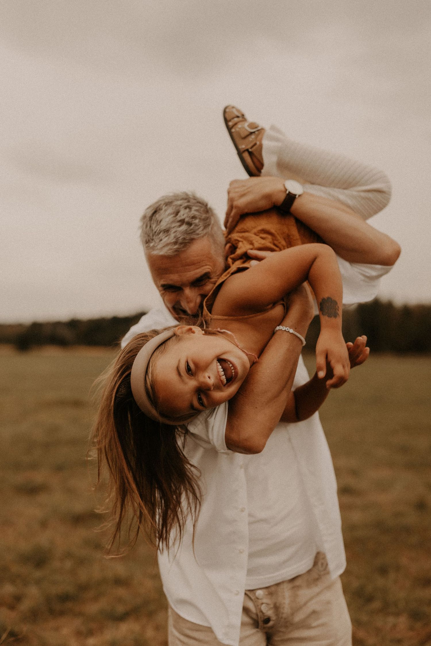 A man playfully lifts a laughing child upside down in a field, both wearing white, sharing a joyful outdoor moment.