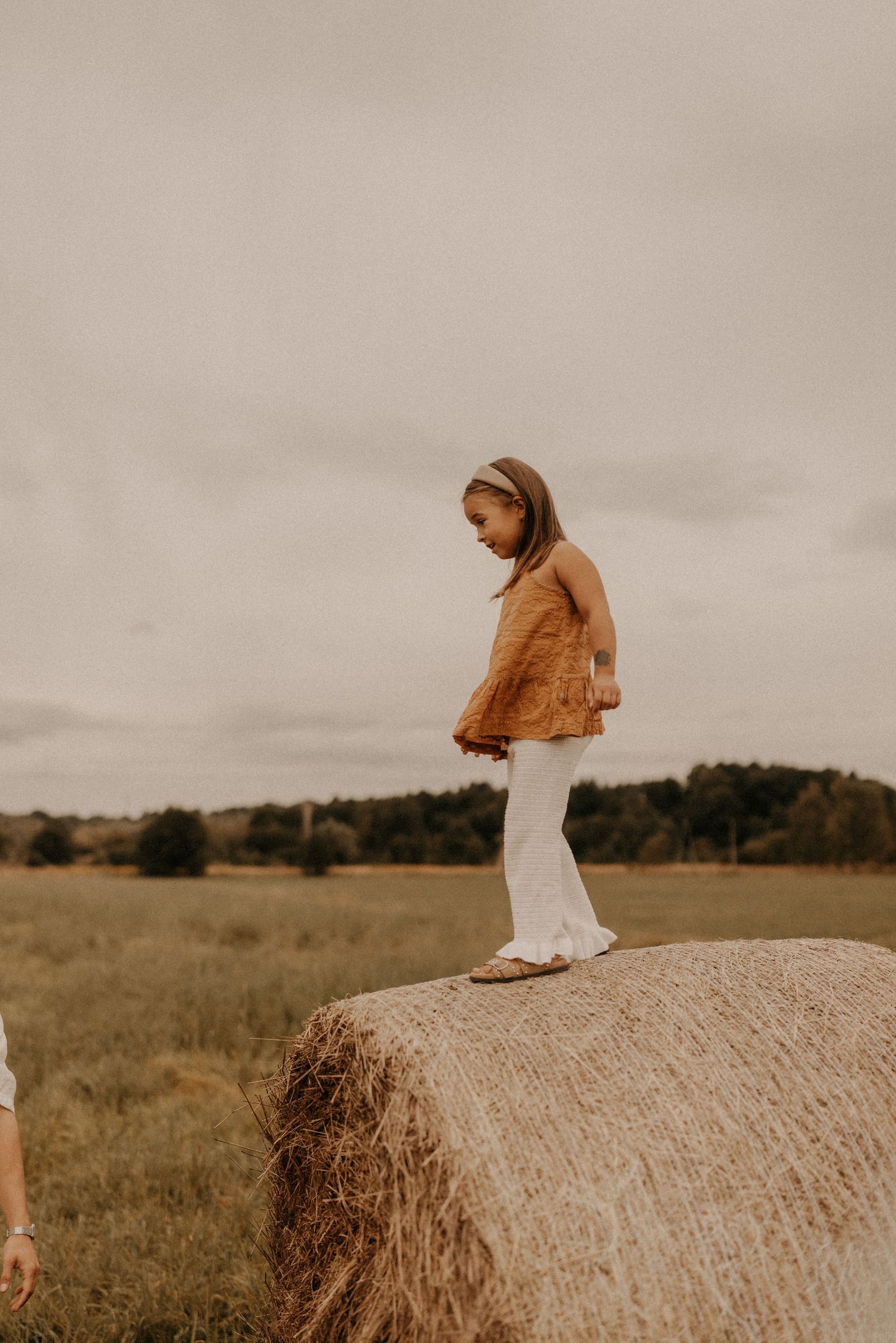 Young girl in brown top and white pants standing on a hay bale in a rural field on an overcast day.