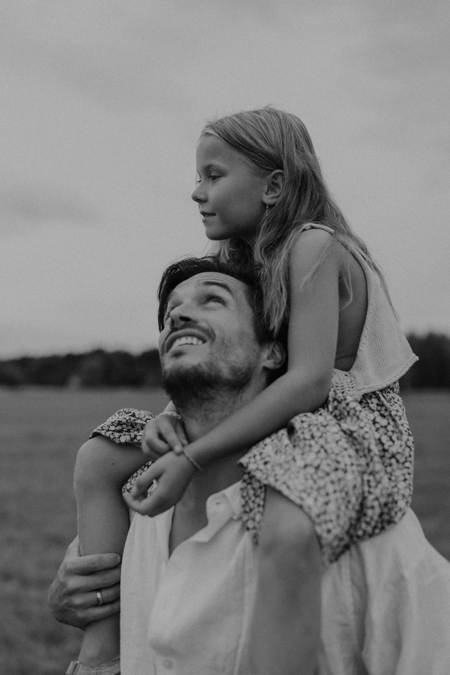 Black and white photo of a smiling father carrying his young daughter on his shoulders in an outdoor field.
