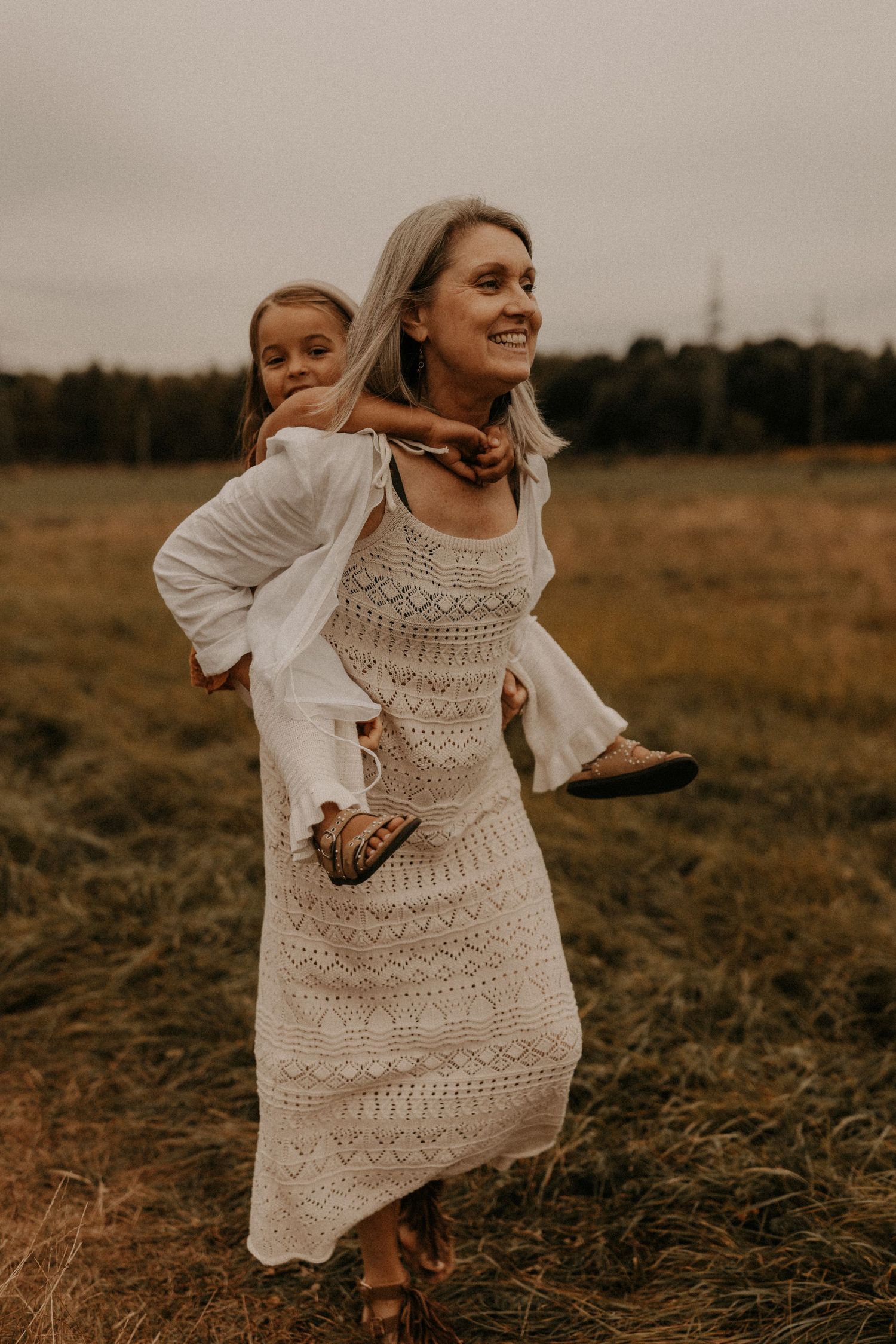 A woman in a white lace dress gives a young girl a piggyback ride in an open field.