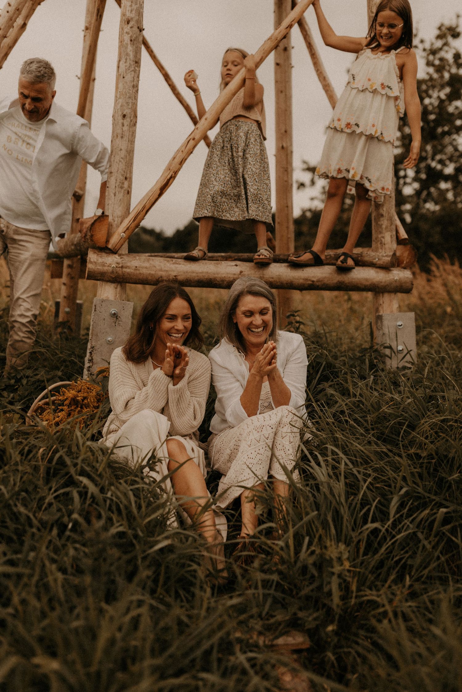 Two women laugh in a field while children play on a rustic wooden structure and a man looks on.