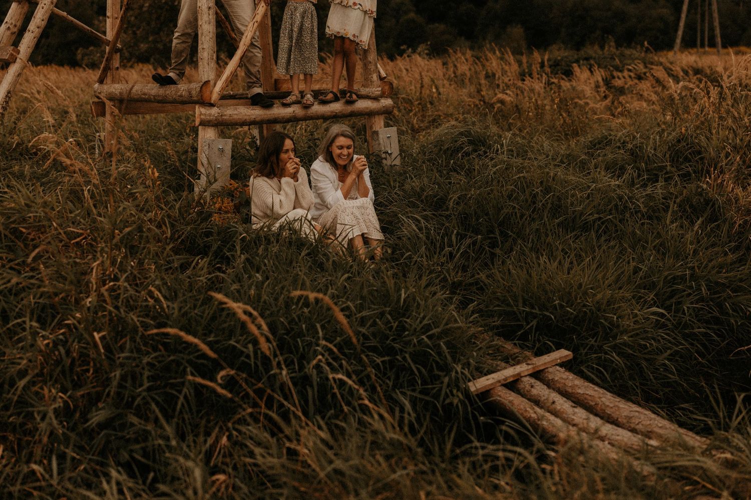 Two women sit laughing in tall golden grass beside a rustic wooden bridge during a warm outdoor family photo session.