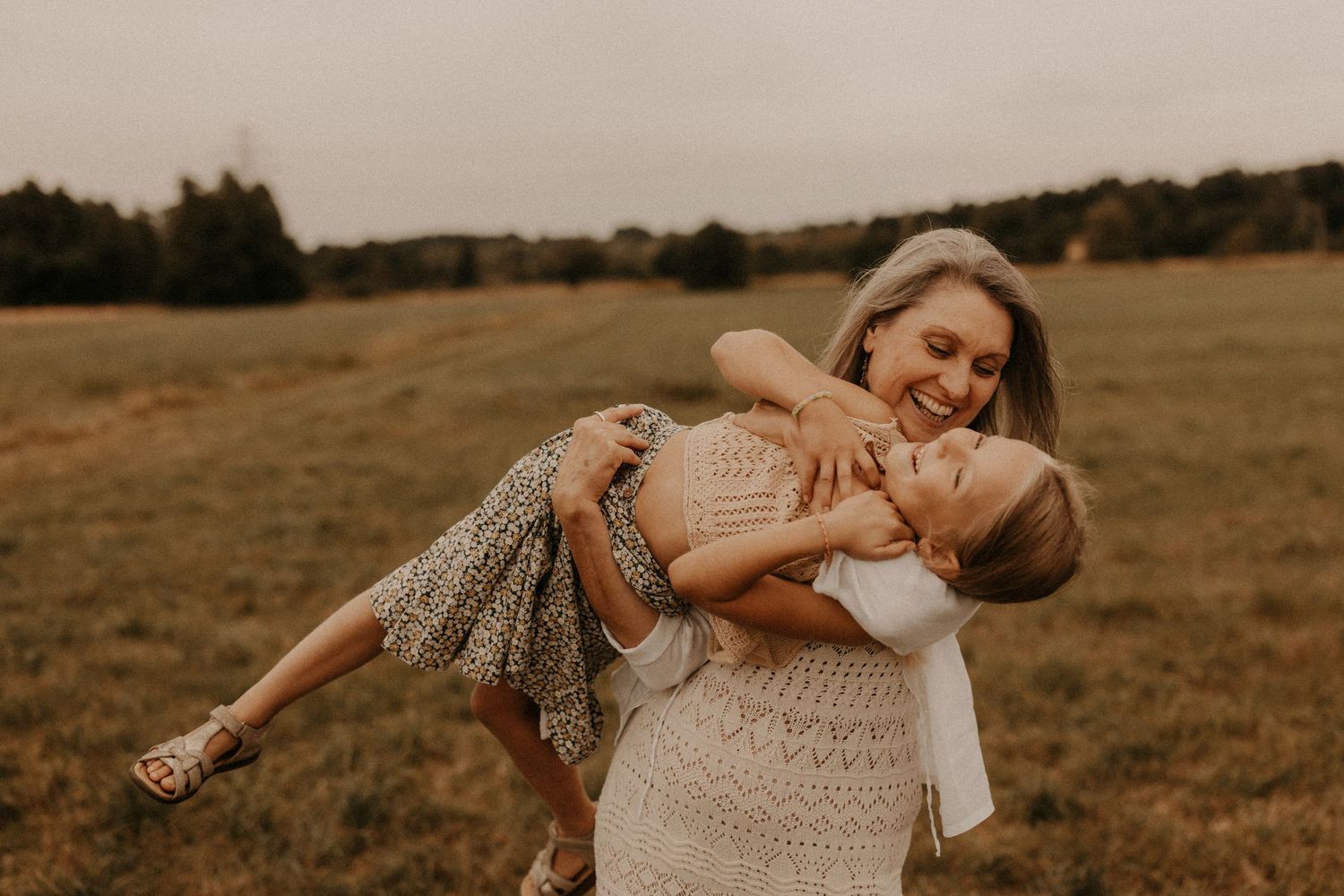 A joyful mother lifts her laughing child in a sunlit green field, both wearing bohemian dresses.