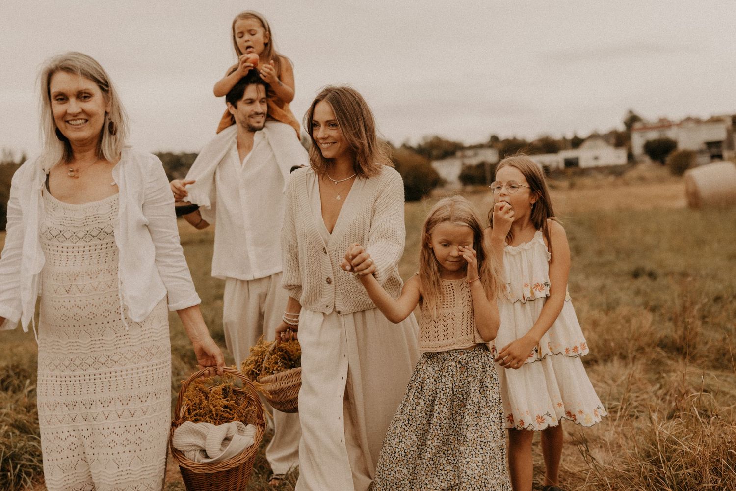 Family walking in a field wearing neutral boho outfits, carrying a basket, with children and two women smiling outdoors.