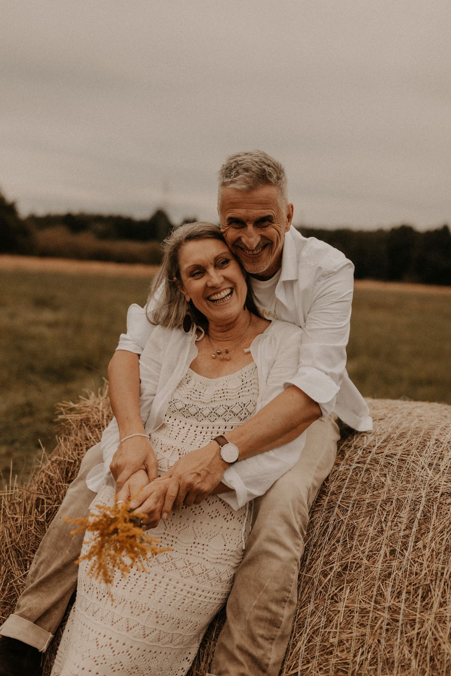 Happy older couple sitting on a hay bale in a field, smiling and embracing during a romantic autumn photoshoot.