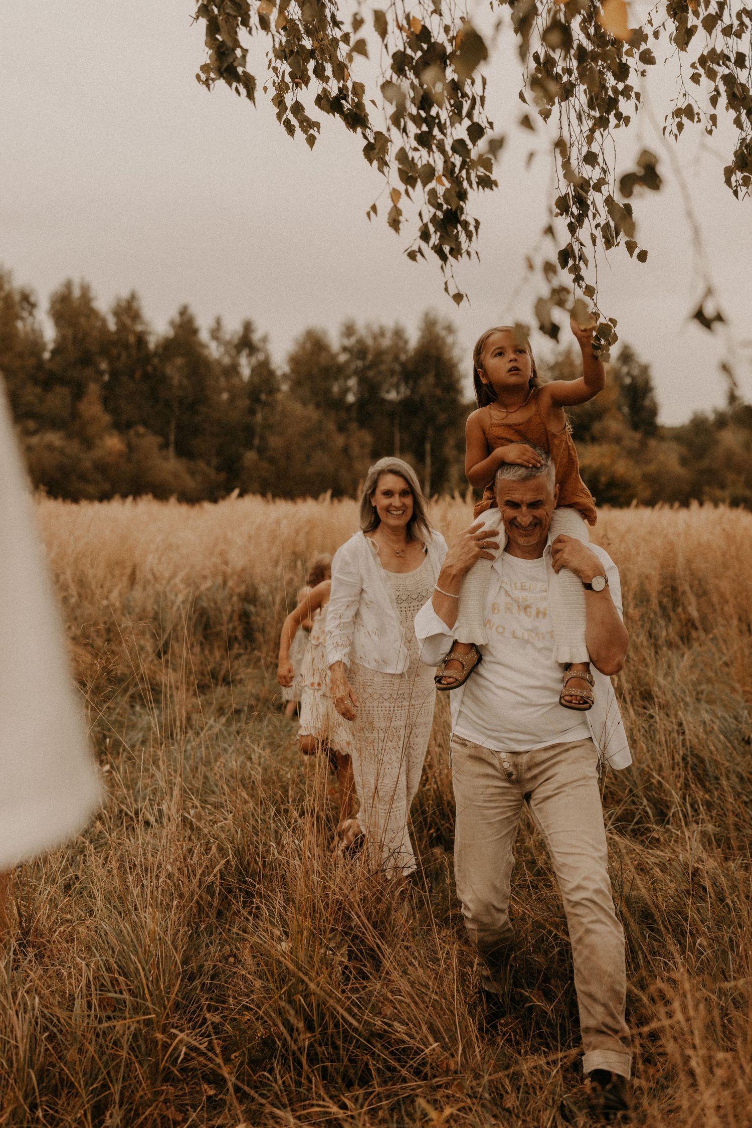 A family walks through a golden field, with a child sitting on dad's shoulders while mom smiles behind them.