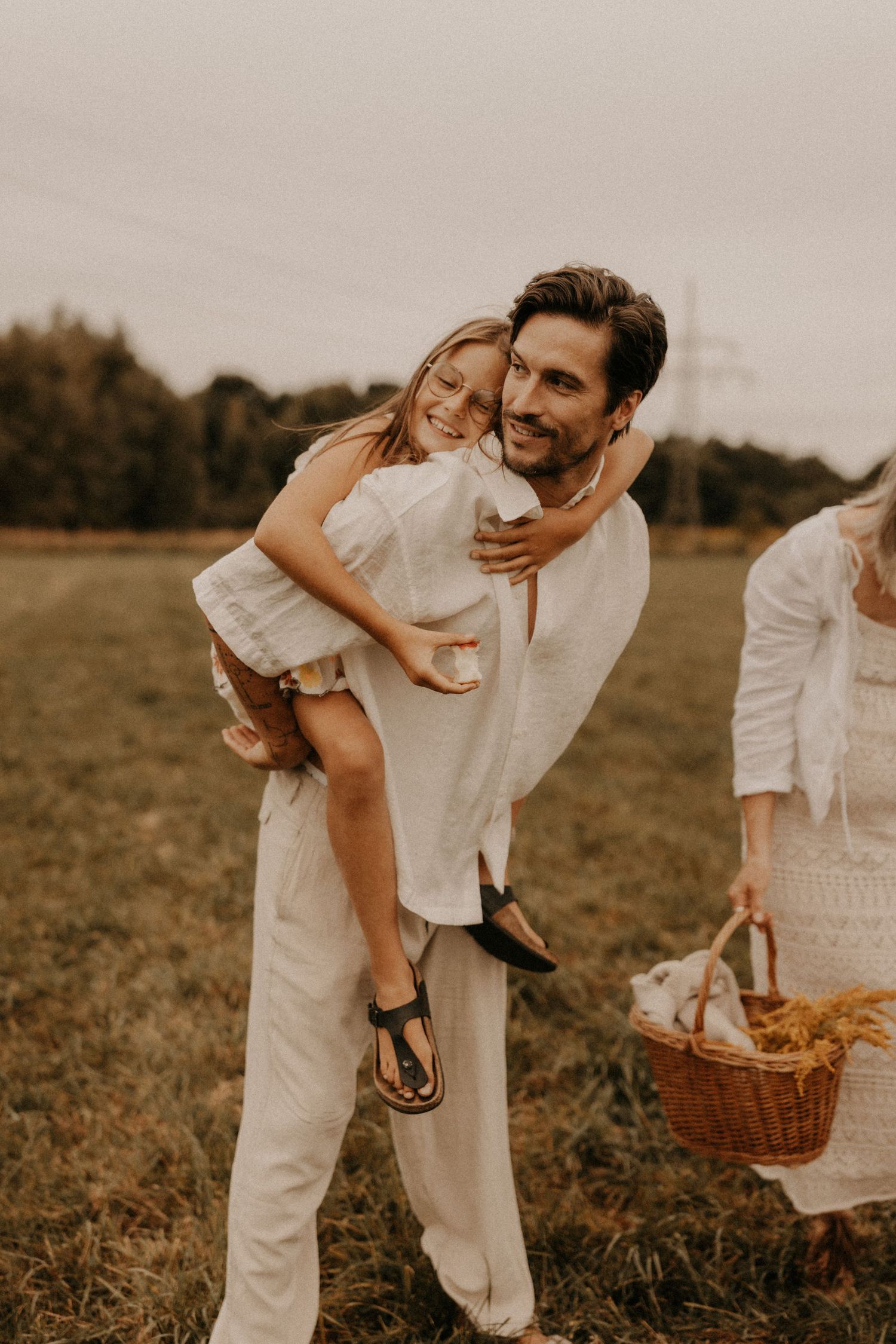 A smiling man in white linen gives a laughing girl a piggyback ride in a field during a family picnic.