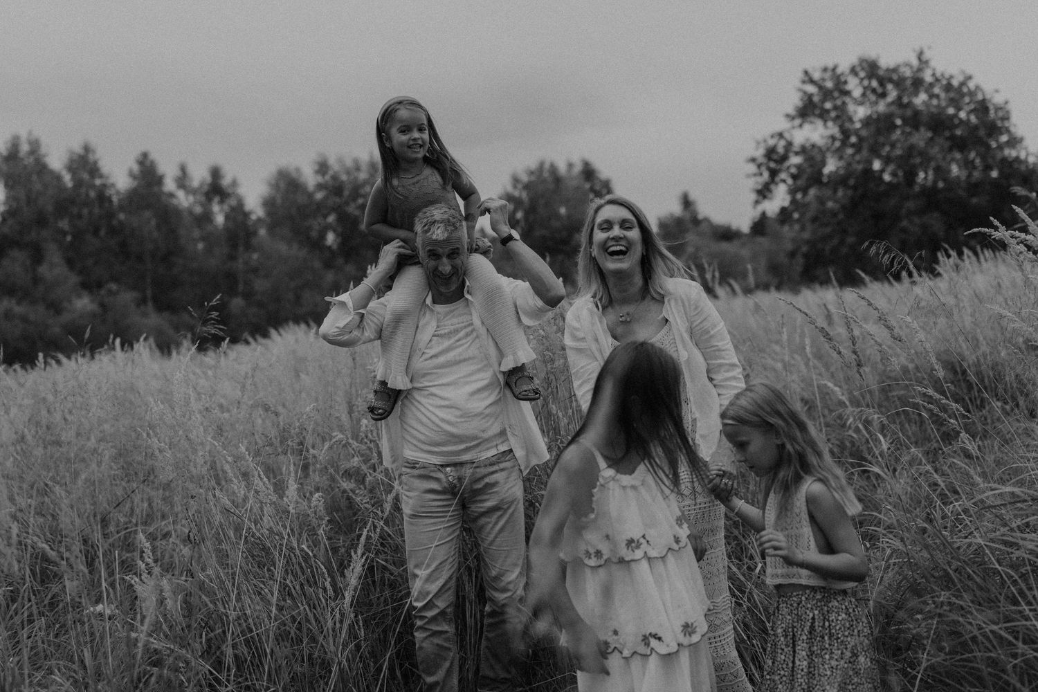 Black and white photo of a joyful family walking through tall grass fields, with children on shoulders and holding hands.
