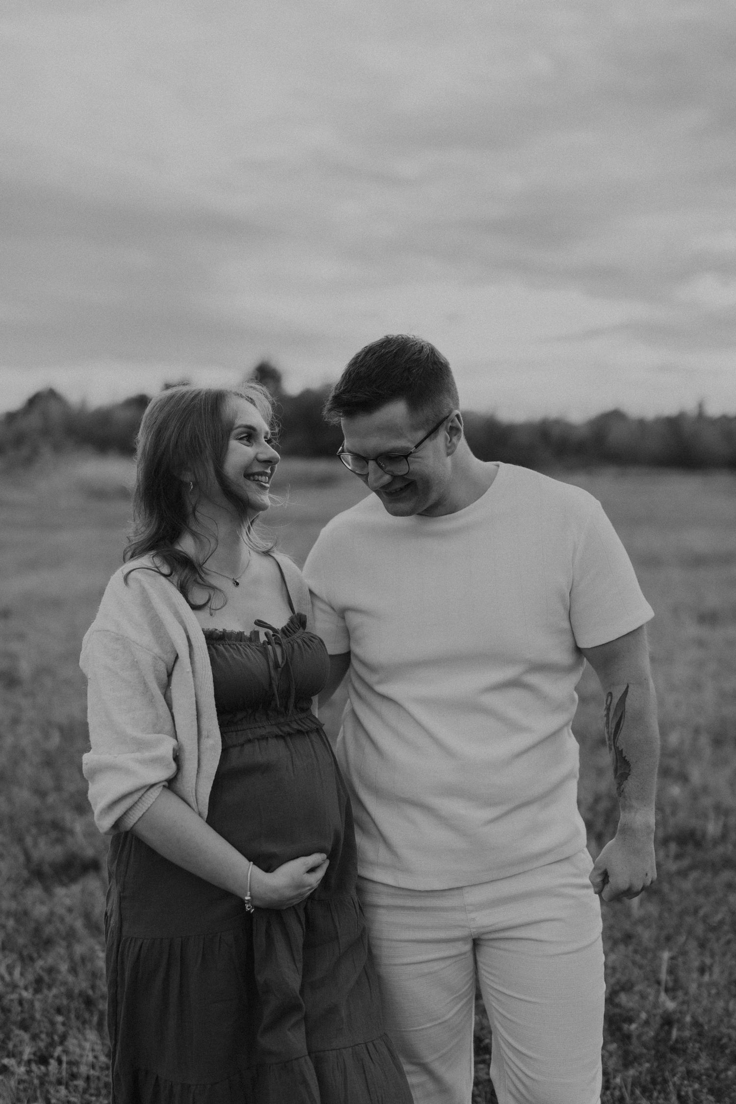 Black and white photo of a pregnant couple standing in a field, the man gazing at the womans baby bump. sesje zdjęciowe Rybnik, Wodzisław Śl