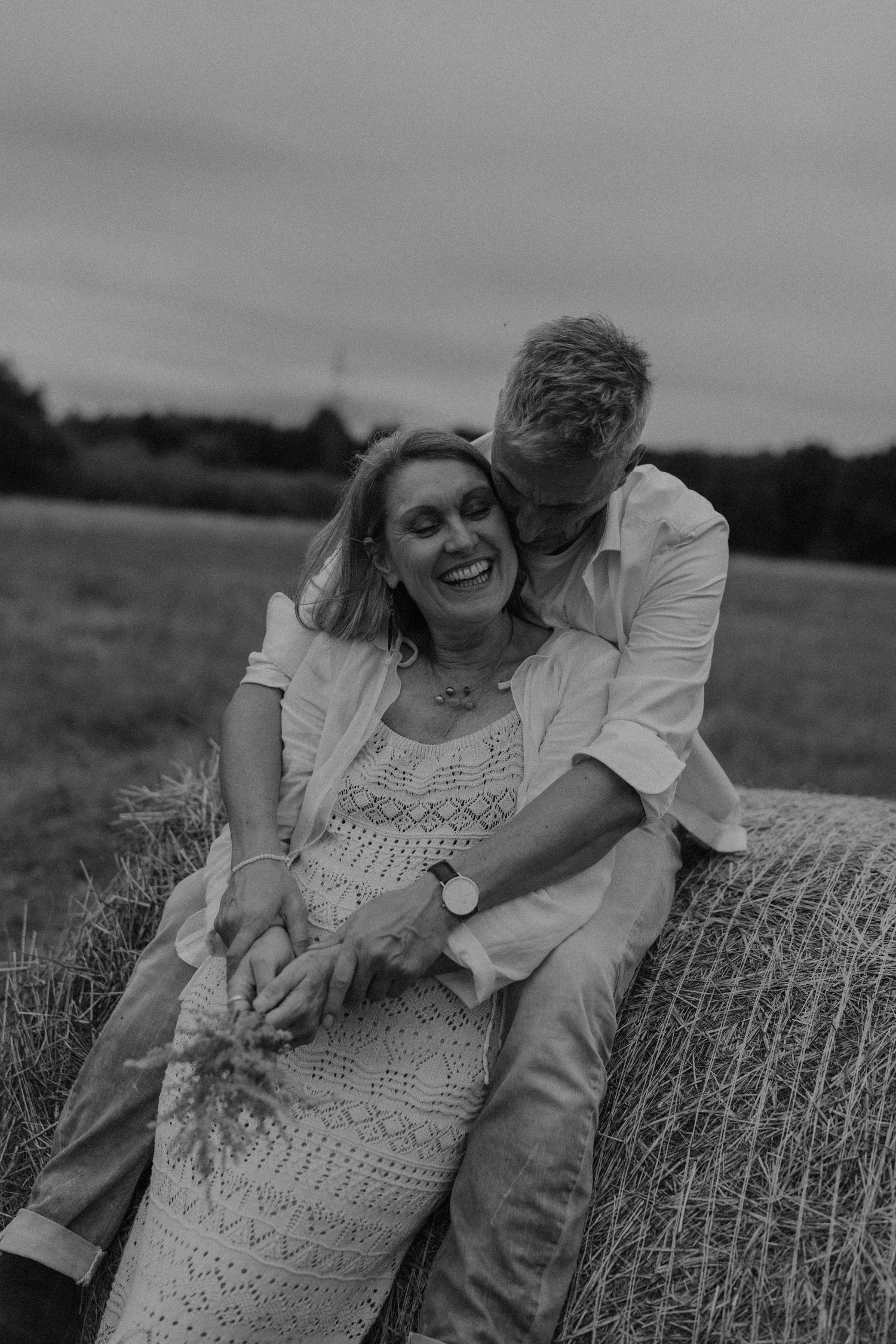 Black and white photo of a smiling couple sitting on a hay bale in a field, man kissing womans cheek.