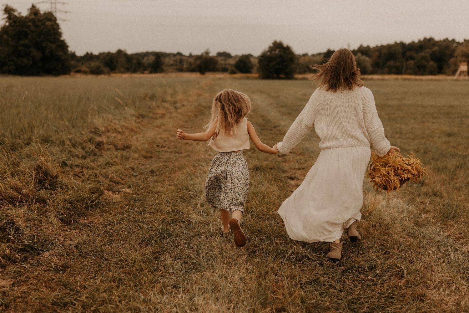 A woman in a white dress and a young girl run hand-in-hand through a golden meadow, carrying a bouquet of yellow flowers.