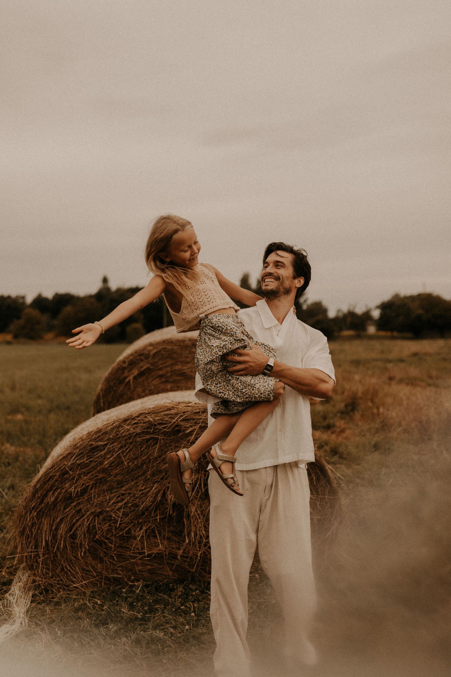 A smiling man lifts a young girl in a floral dress near a hay bale in an open field.