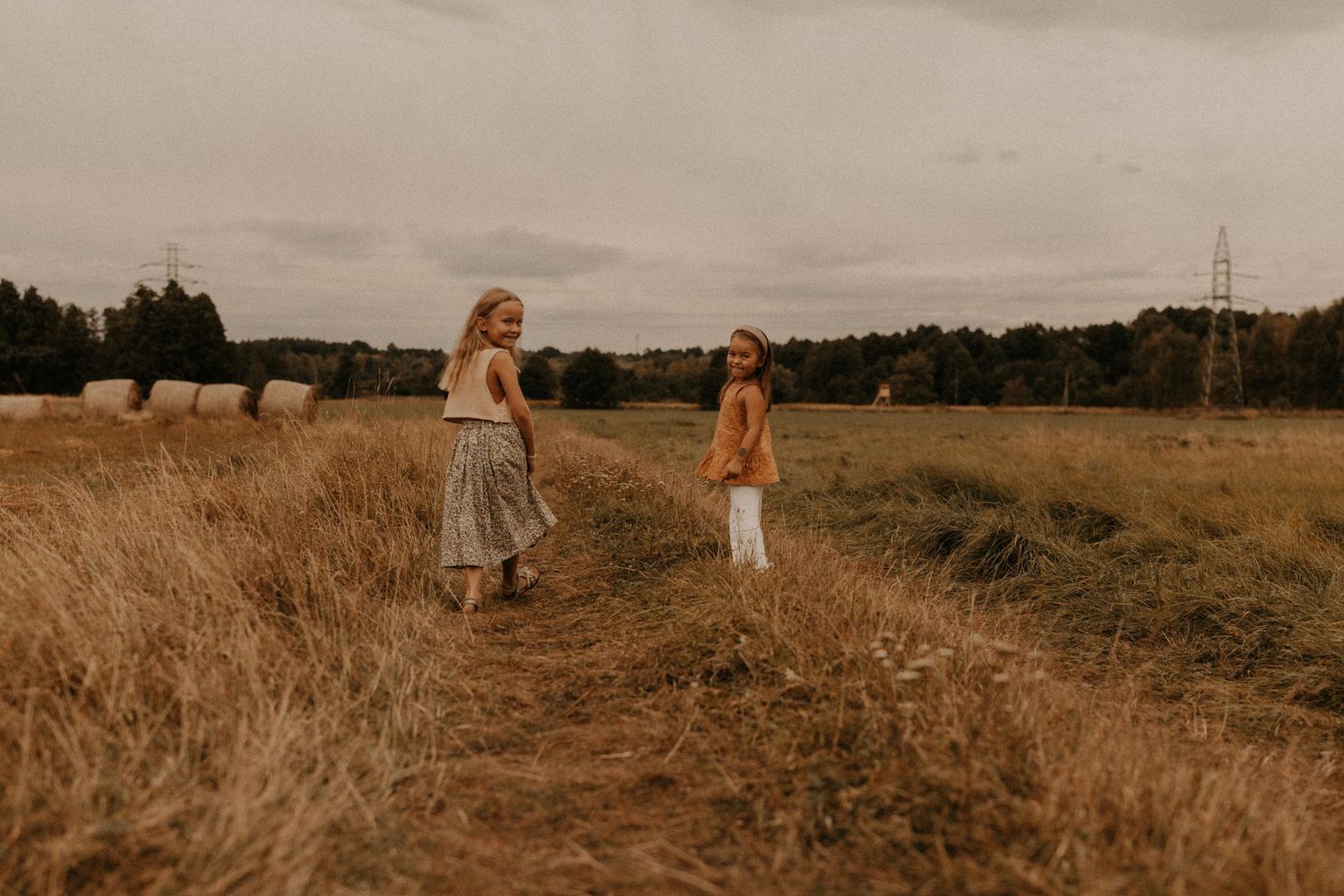 Two young girls walking through a golden field with hay bales and trees in the background on an overcast day.