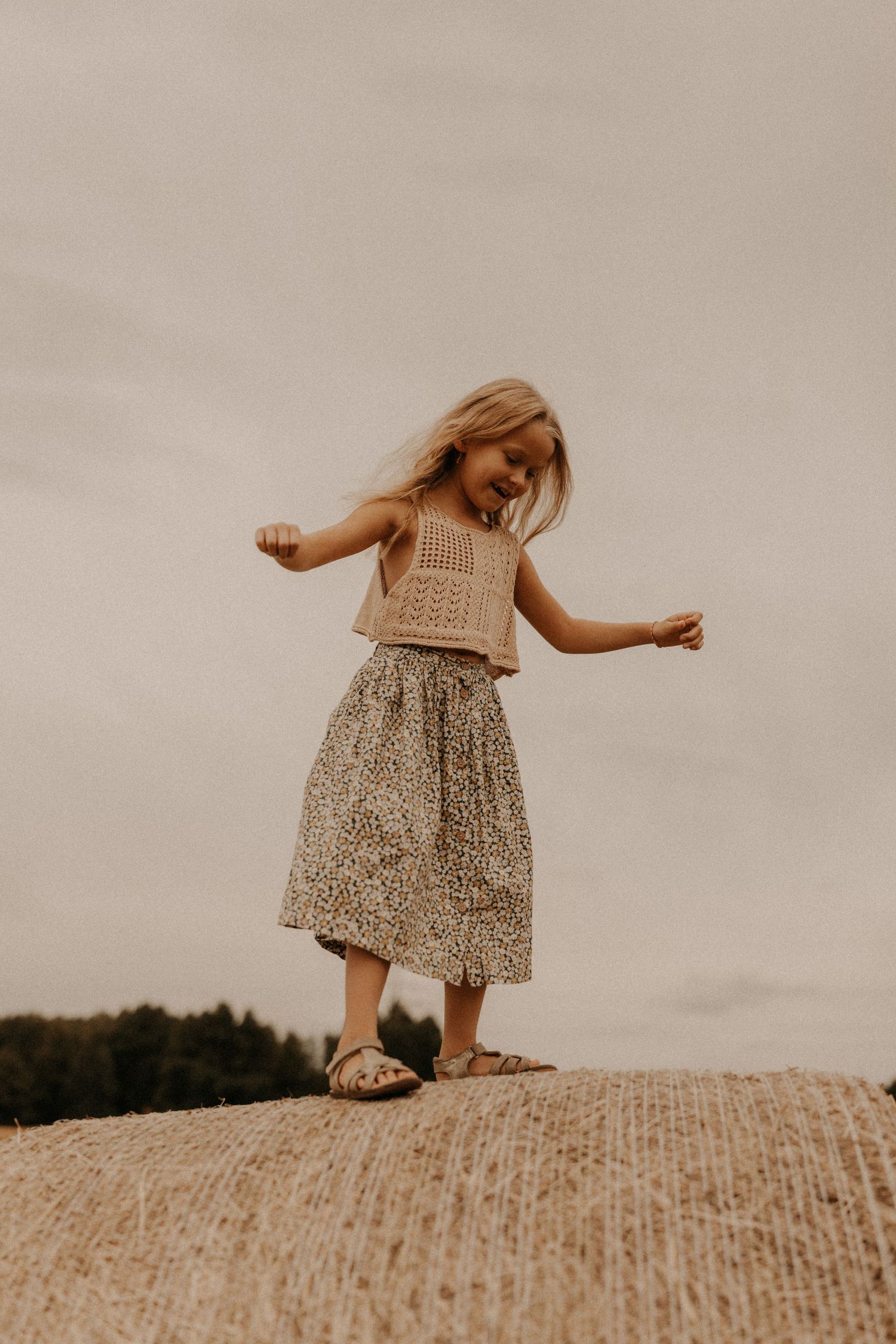 Young girl balancing on hay bale wearing crochet top and floral midi skirt with sandals outdoors.