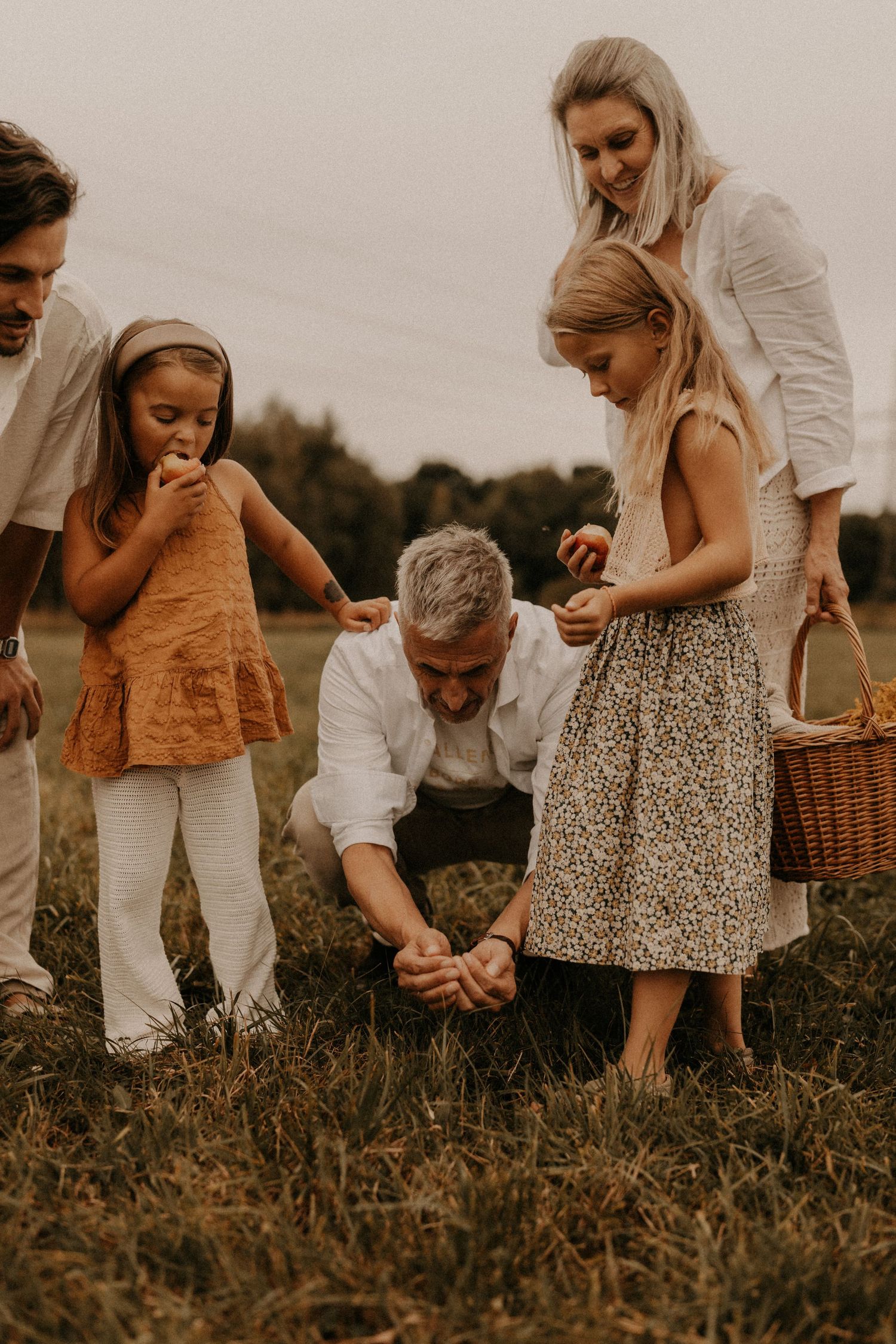 Family of five enjoying a peaceful outdoor gathering in a field, with children holding a basket and apple on a cloudy day.