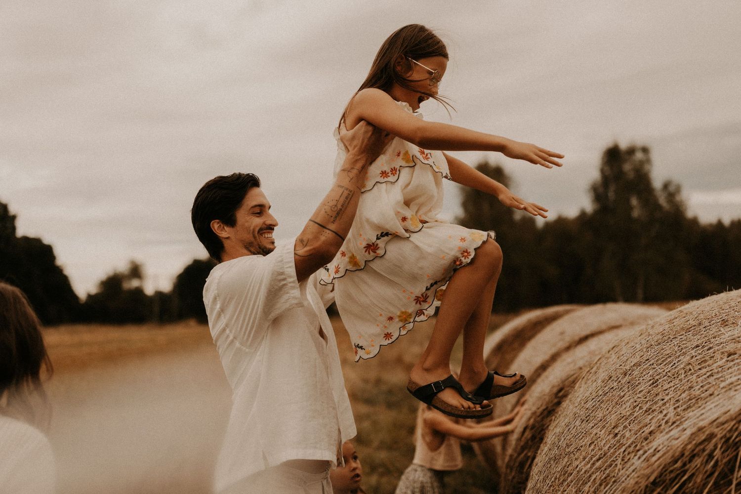 A smiling man lifts a young girl in a floral dress outdoors near hay bales in a rustic field setting.