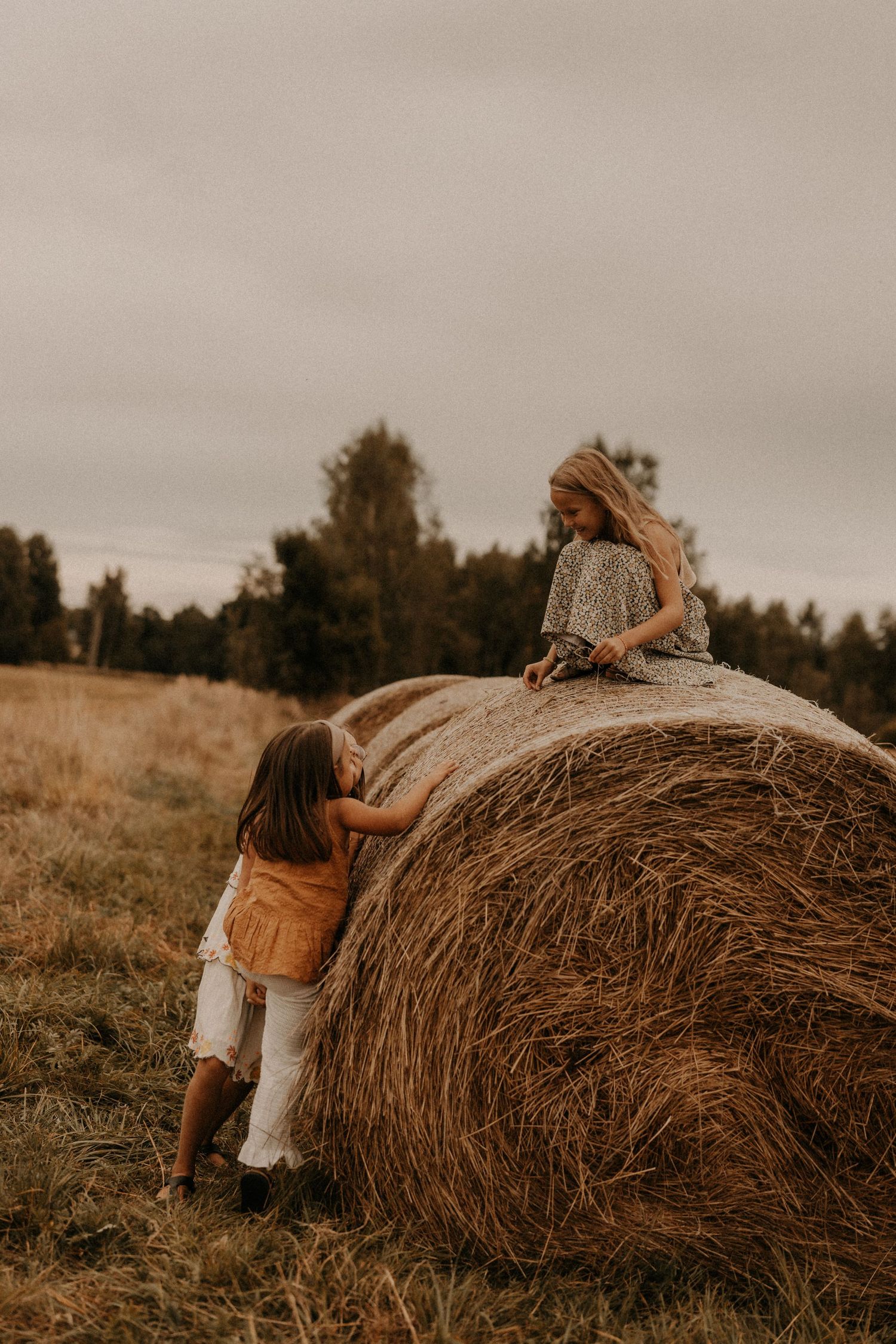 Two girls playing on a large round hay bale in an open field with trees and overcast sky in the background.