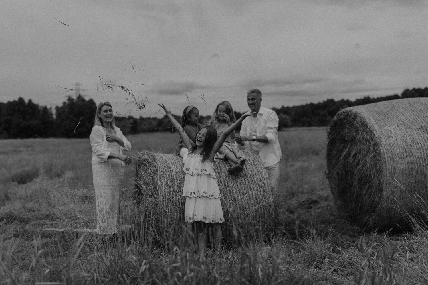 Black and white photo of a joyful family in traditional attire celebrating near hay bales in a rustic field.