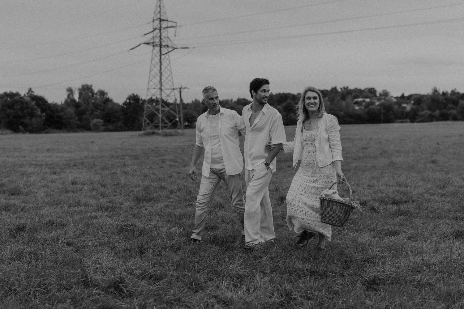 Three people walking through a green field near a power line tower in a black and white outdoor photo.