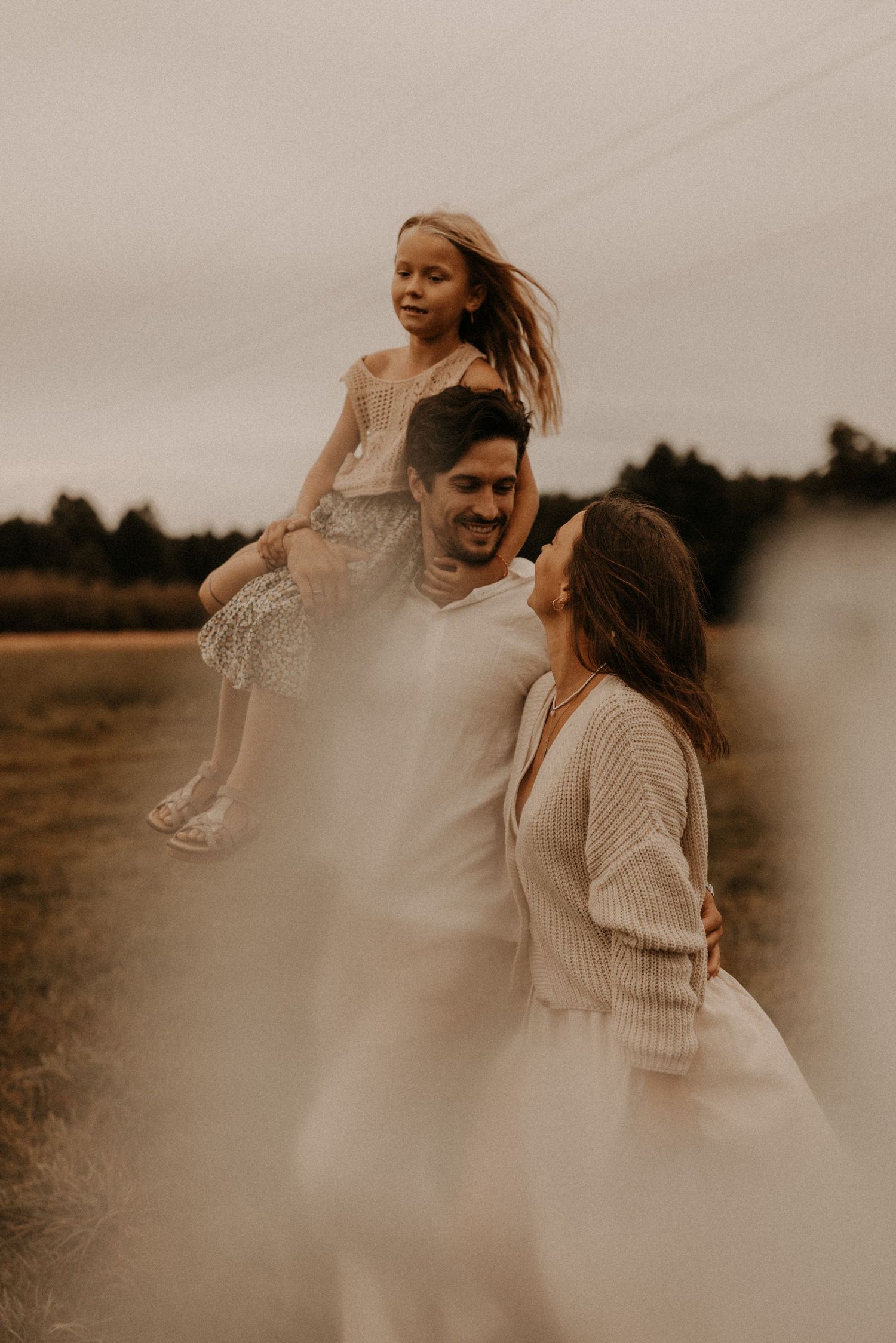 A happy family of three poses outdoors, with dad lifting daughter on shoulders while mom smiles in a golden field.